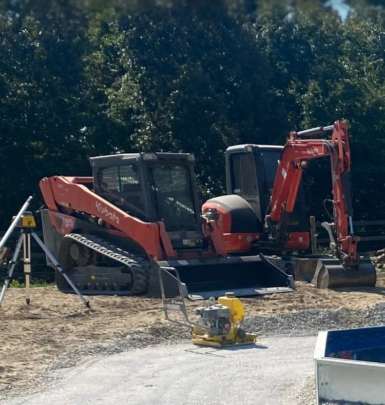 Construction site with a red Kubota excavator and a compaction machine on a dirt road, with trees in the background.