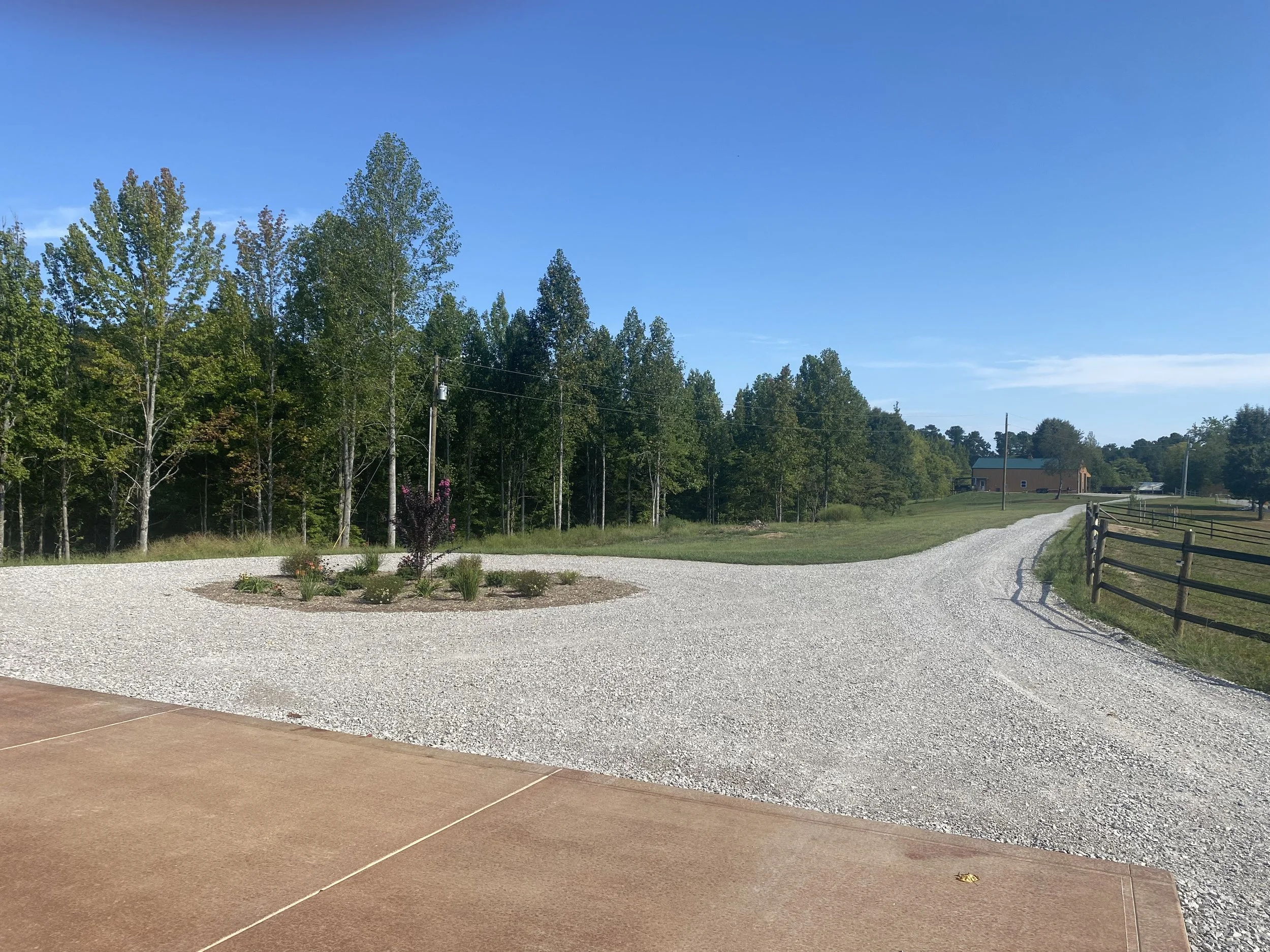 A dirt and gravel driveway curves through a rural property with a house in the distance, surrounded by trees against a blue sky.