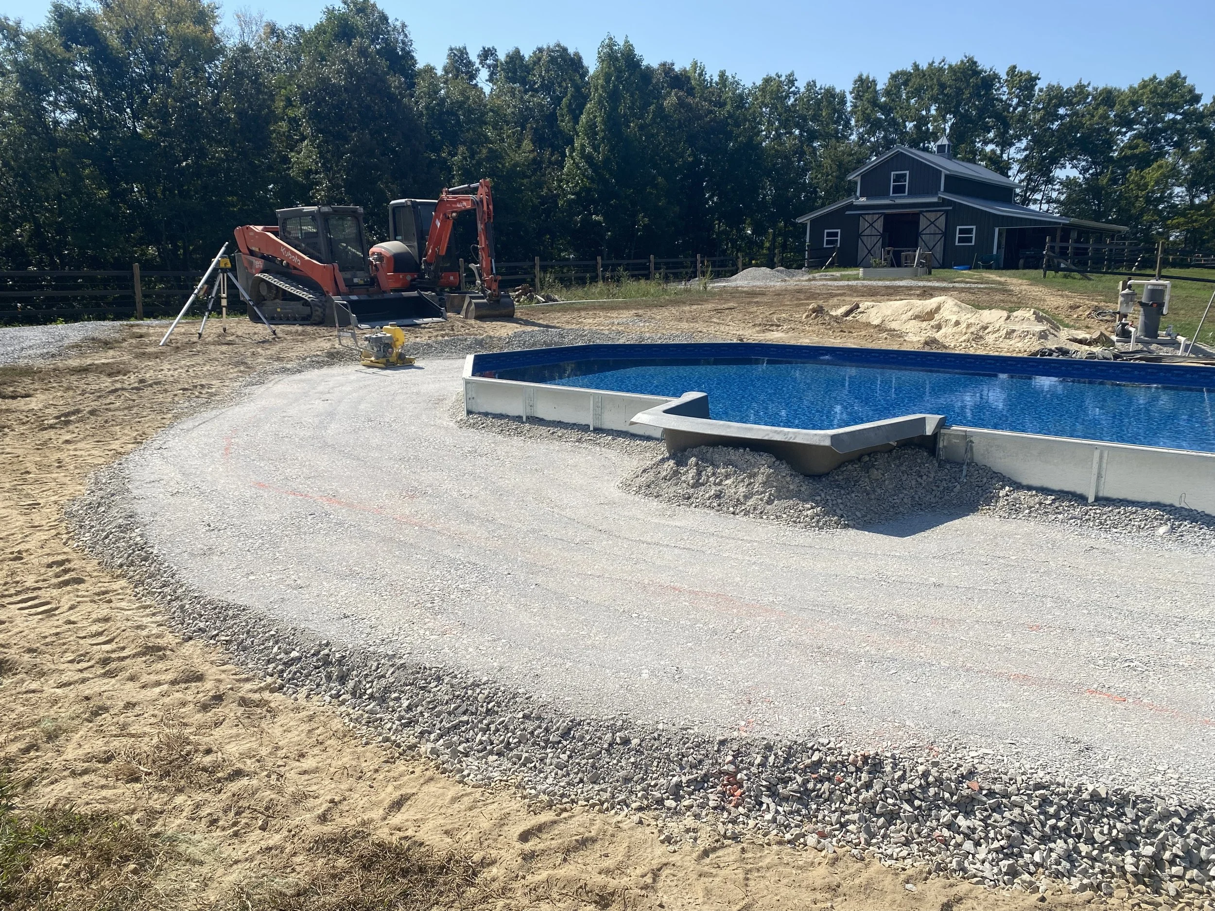 Construction site with a small excavator, gravel, and a swimming pool in front of a blue barn-style house with trees in the background.