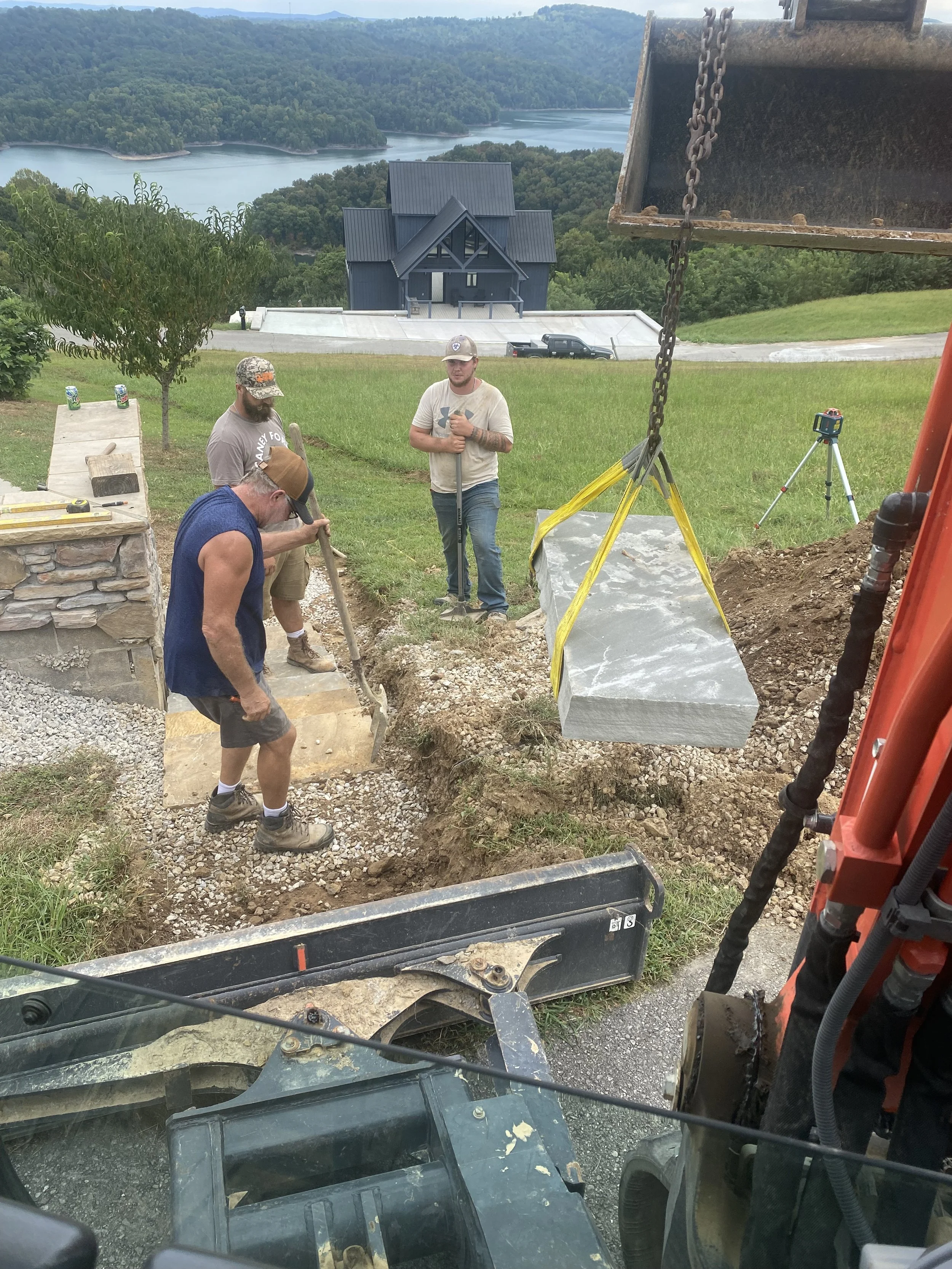 Three workers are outdoors in a yard, working with a large piece of stone. One man is shoveling gravel, another is standing while holding a tool, and a third is working on the ground. There are trees, a house, and a lake with hills in the background.