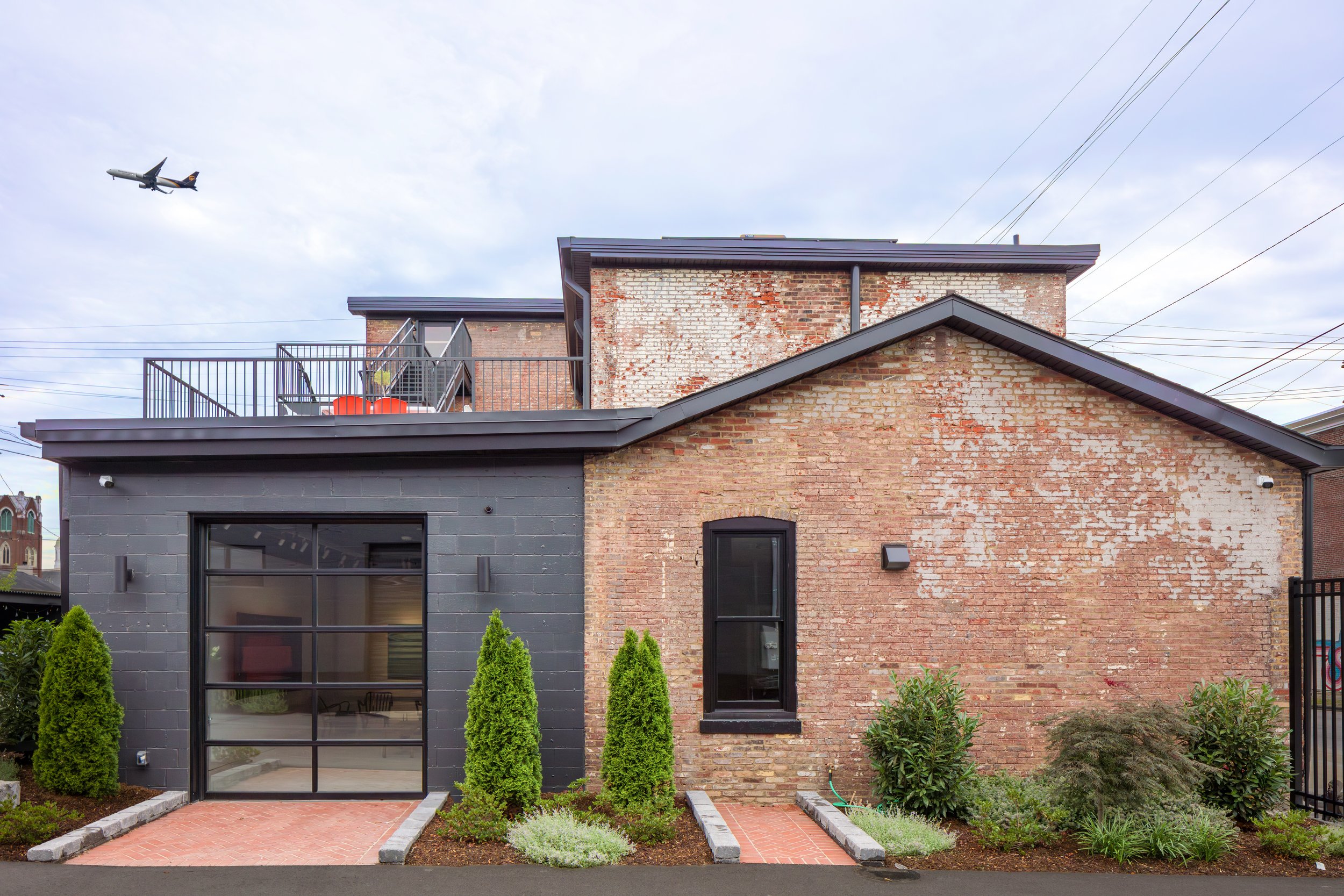 A modern brick and gray house with a balcony, surrounded by a landscaped garden with small trees and shrubs, and an airplane flying in the cloudy sky above.