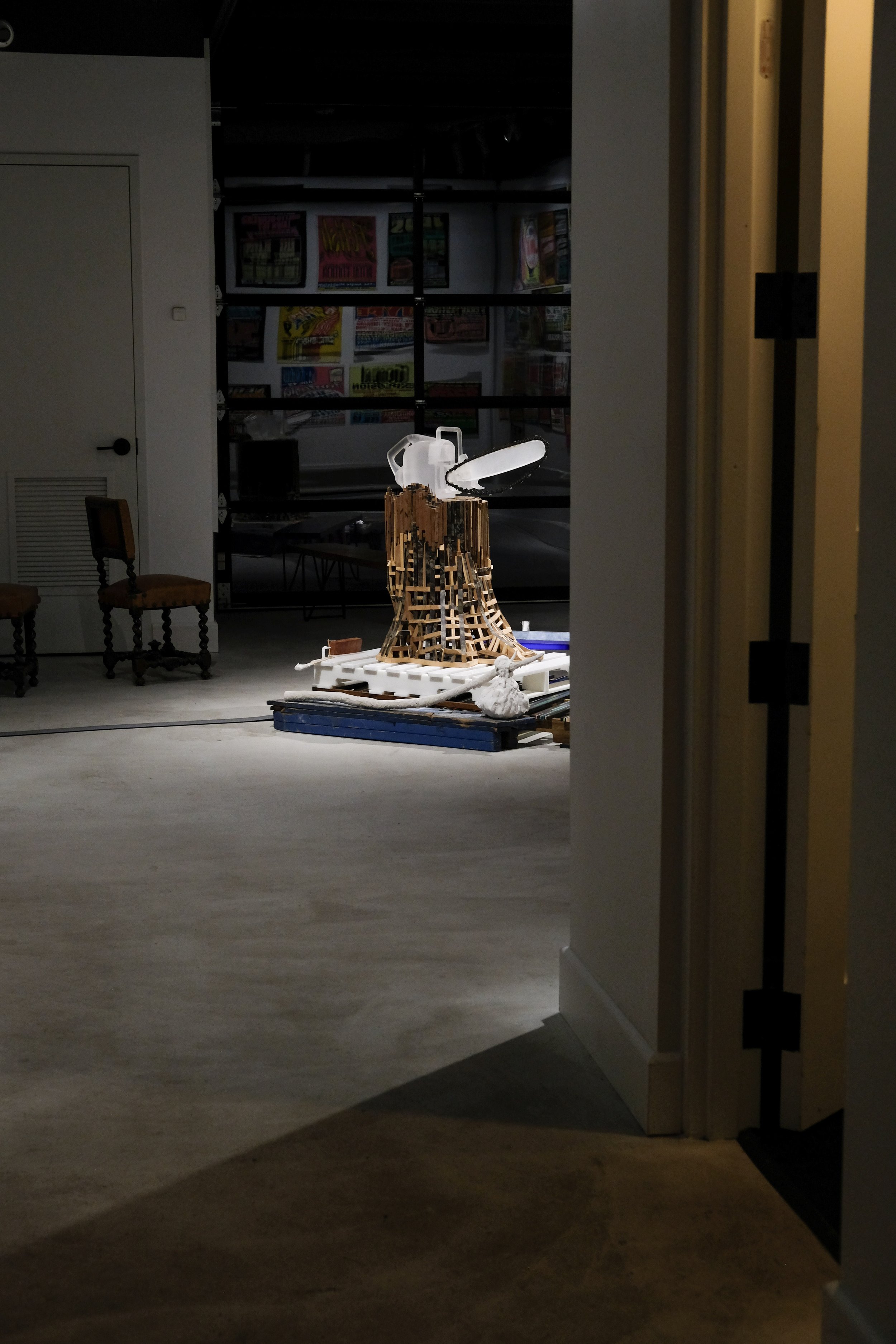 An art installation featuring a wooden sculpture with a saw on top, situated on a platform in a gallery space with posters on the wall in the background.