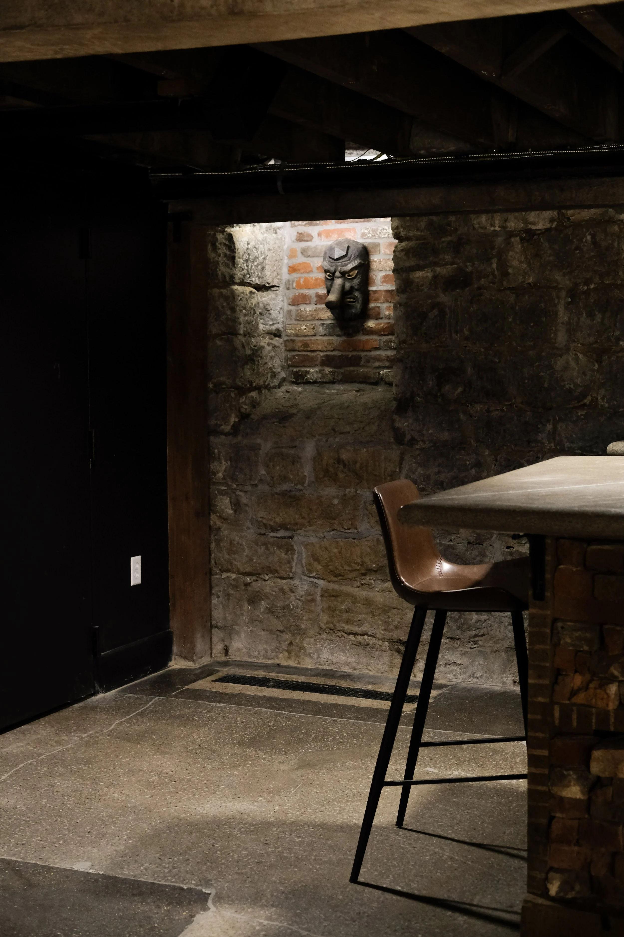 Interior of a room with rustic brick and stone walls, wooden and metal furniture, and a carved face mask hanging on the wall.