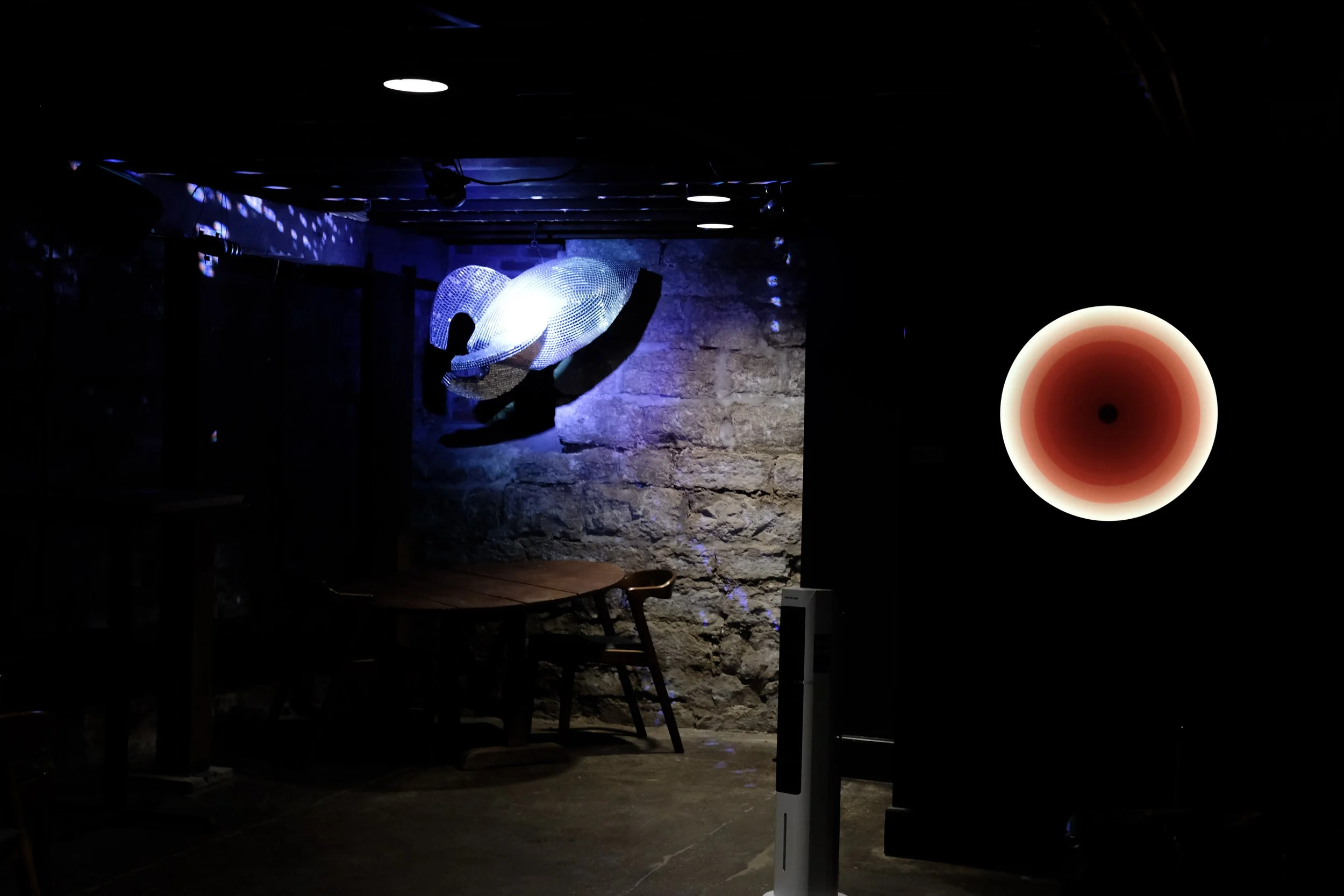 Dimly lit indoor space with a stone wall, seating area, illuminated abstract art installation, and a circular light fixture on the wall.