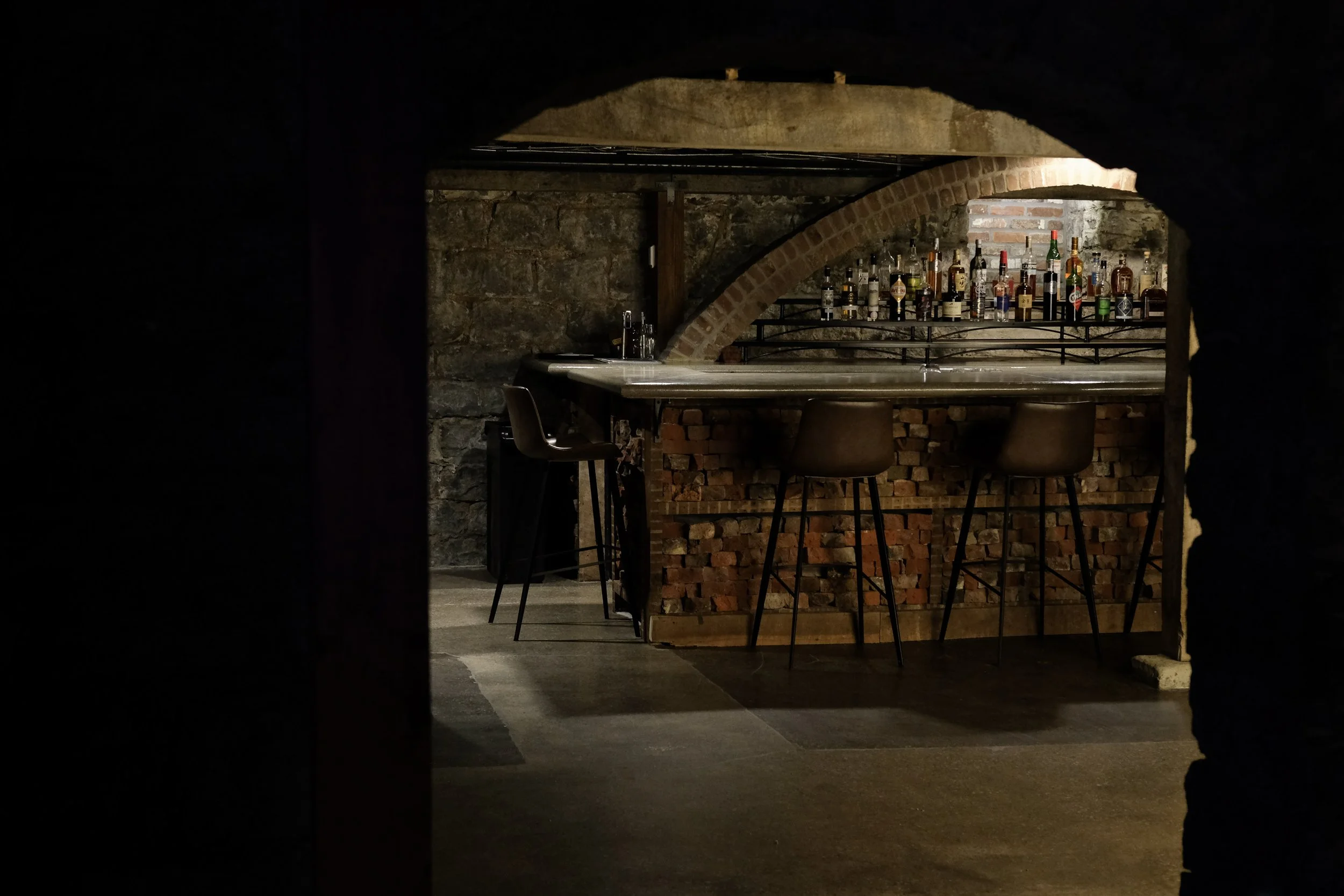 View of a bar with bottles of alcohol on shelves, exposed brick and stone walls, and bar stools.