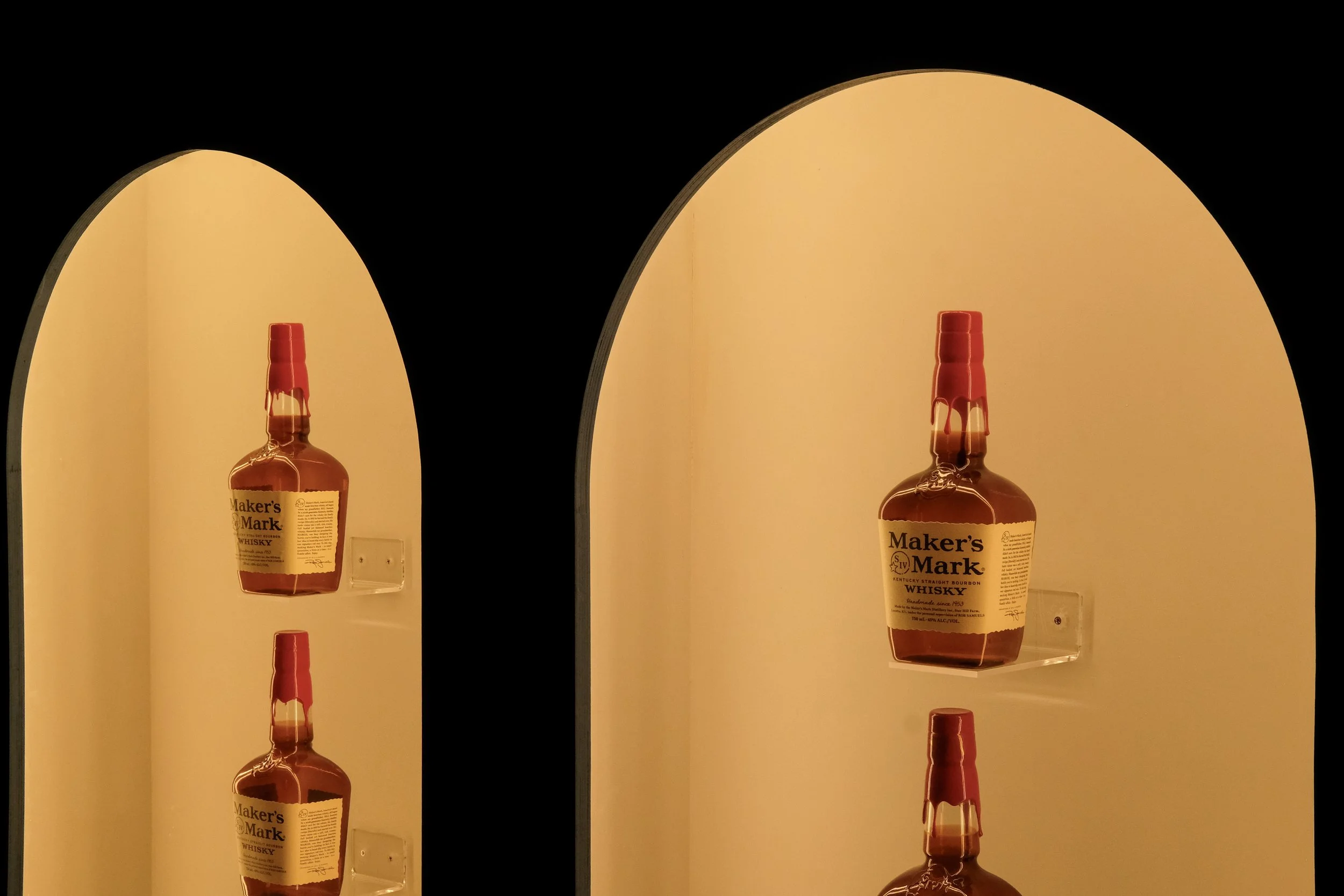 Four bottles of Maker's Mark whiskey displayed on illuminated shelves, with the focus on the middle bottles and part of the other two bottles visible in a dimly lit setting.