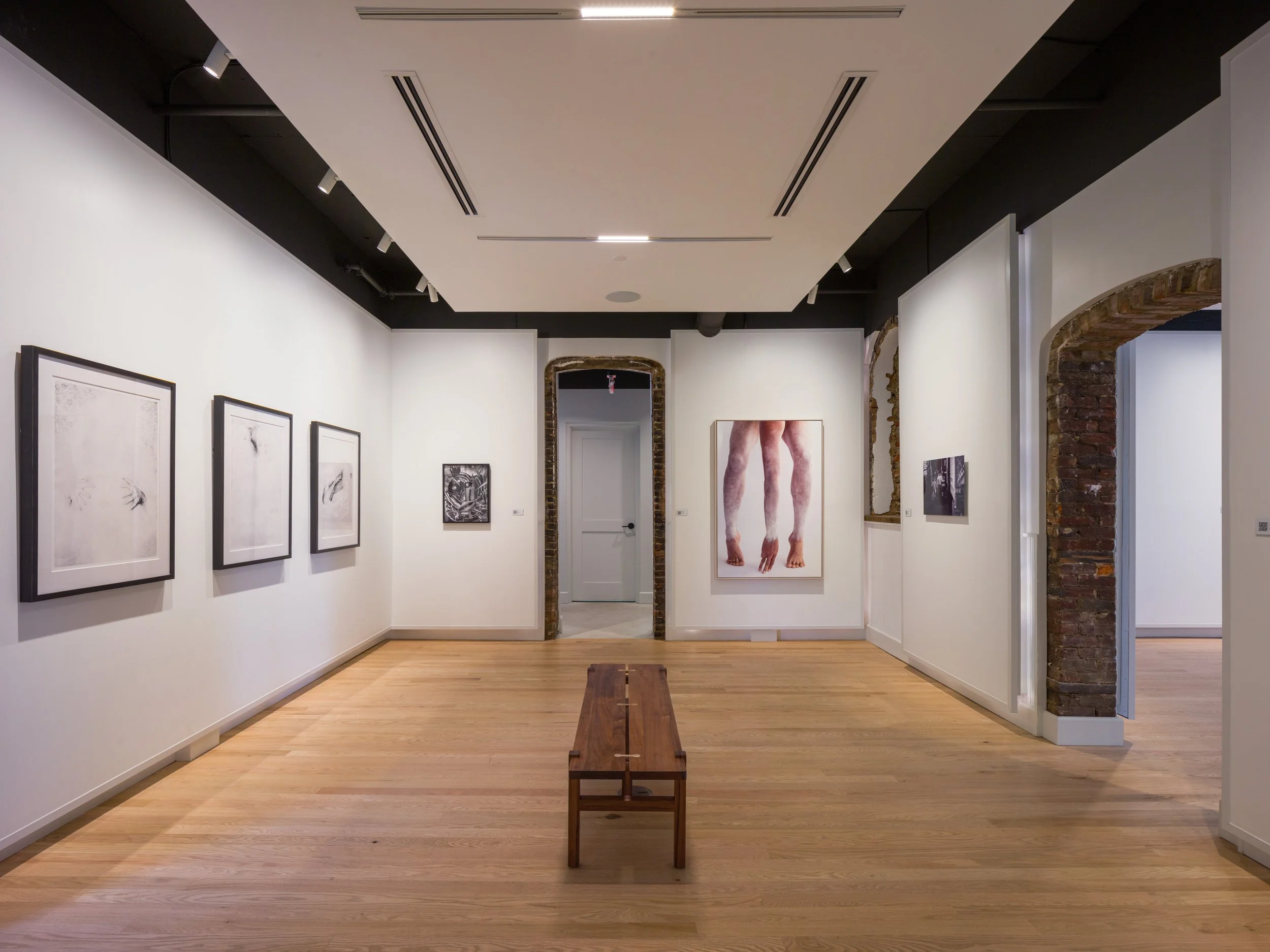 An art gallery with white walls and wooden floors, featuring various framed artworks and photographs. A wooden bench is centered in the room, and a large photograph of human legs is visible on the back wall. Exposed brick arches are on the right, and the ceiling has track lighting.