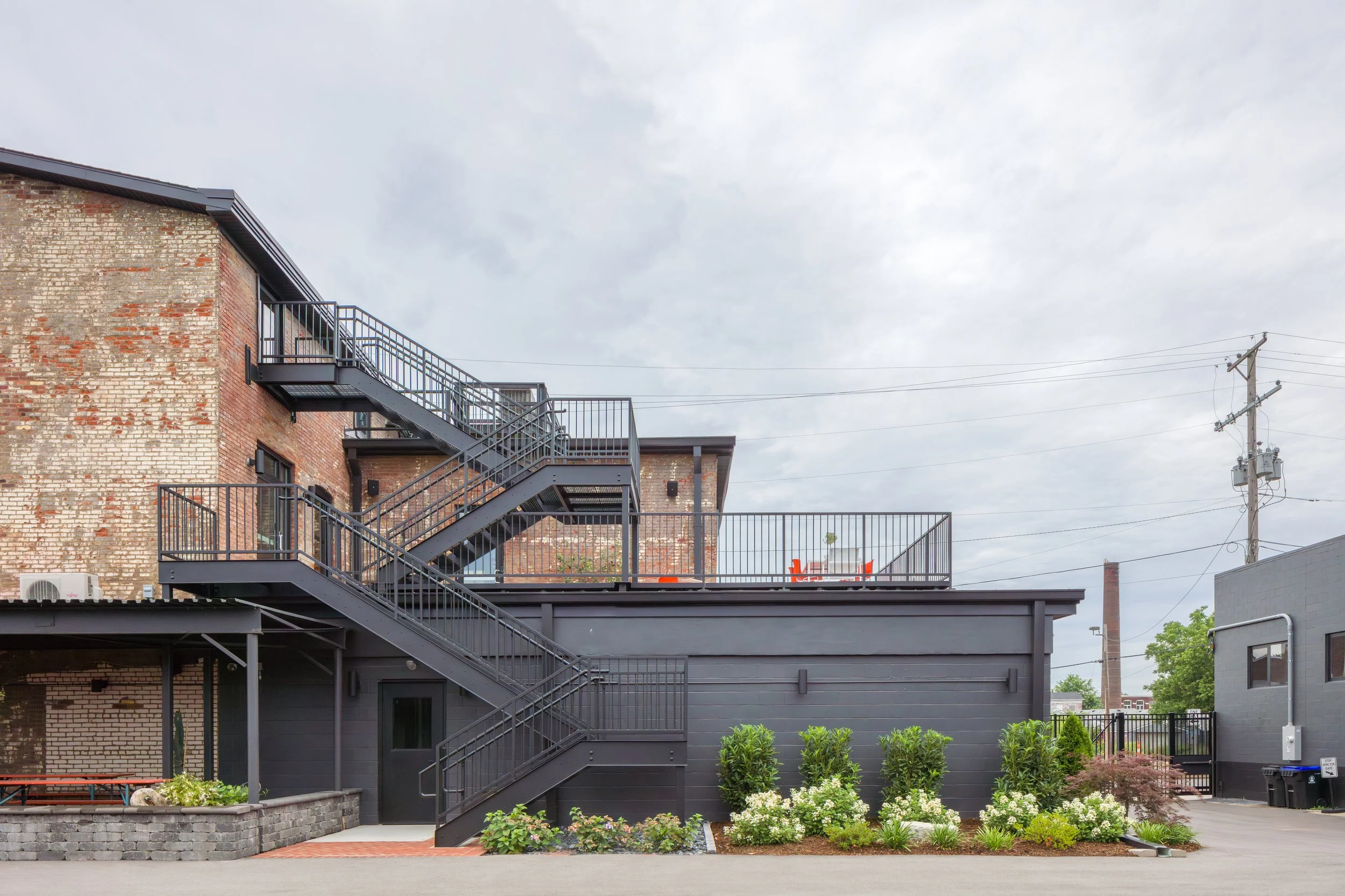 Exterior view of a modern brick building with black metal fire escape stairs and railing, featuring a landscaped area with green shrubs and flowers, under a cloudy sky and utility poles in the background.