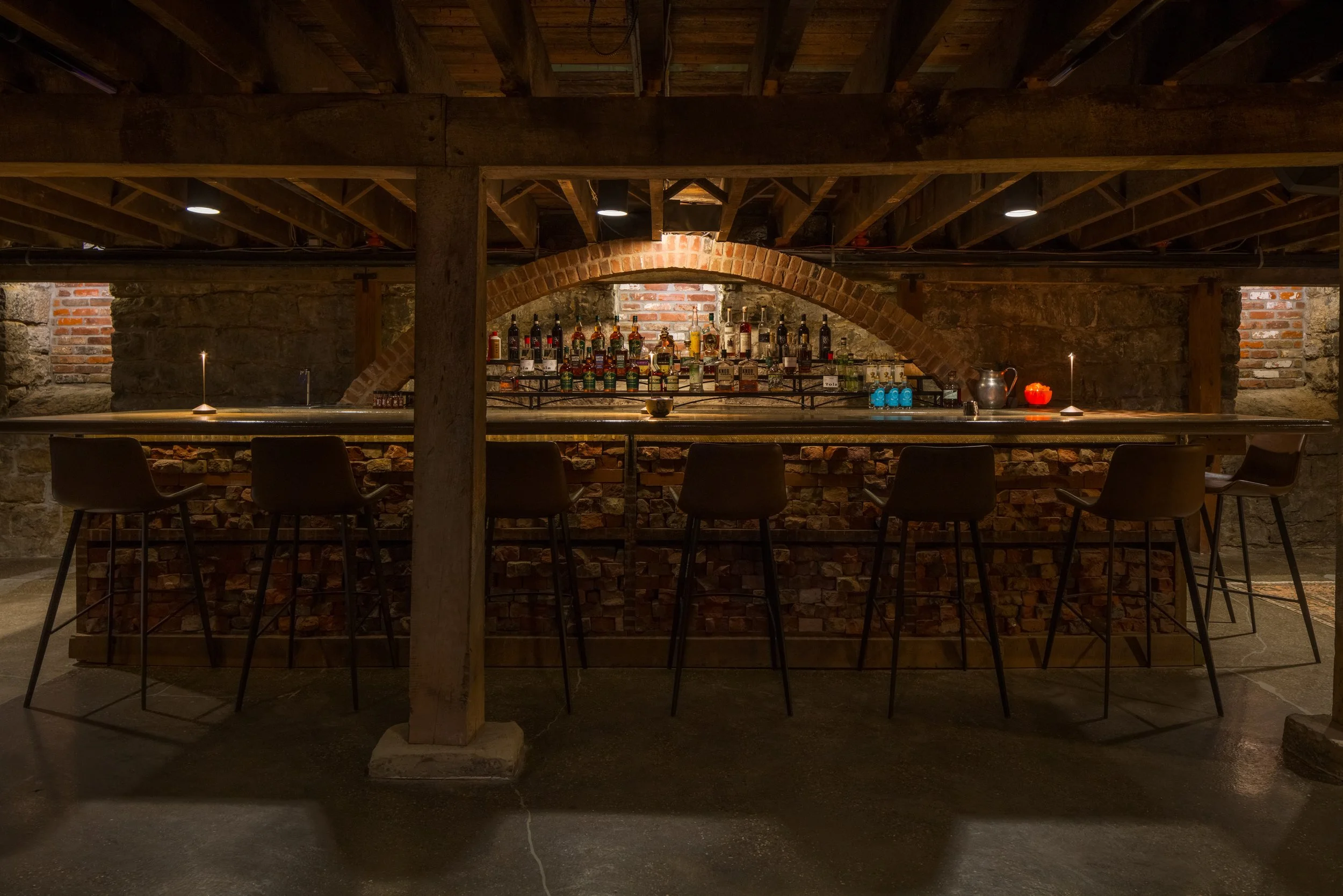 Bar area with bottles on shelves, exposed brick and wood beams, dim lighting, candles, and chairs in a rustic basement or cellar setting.