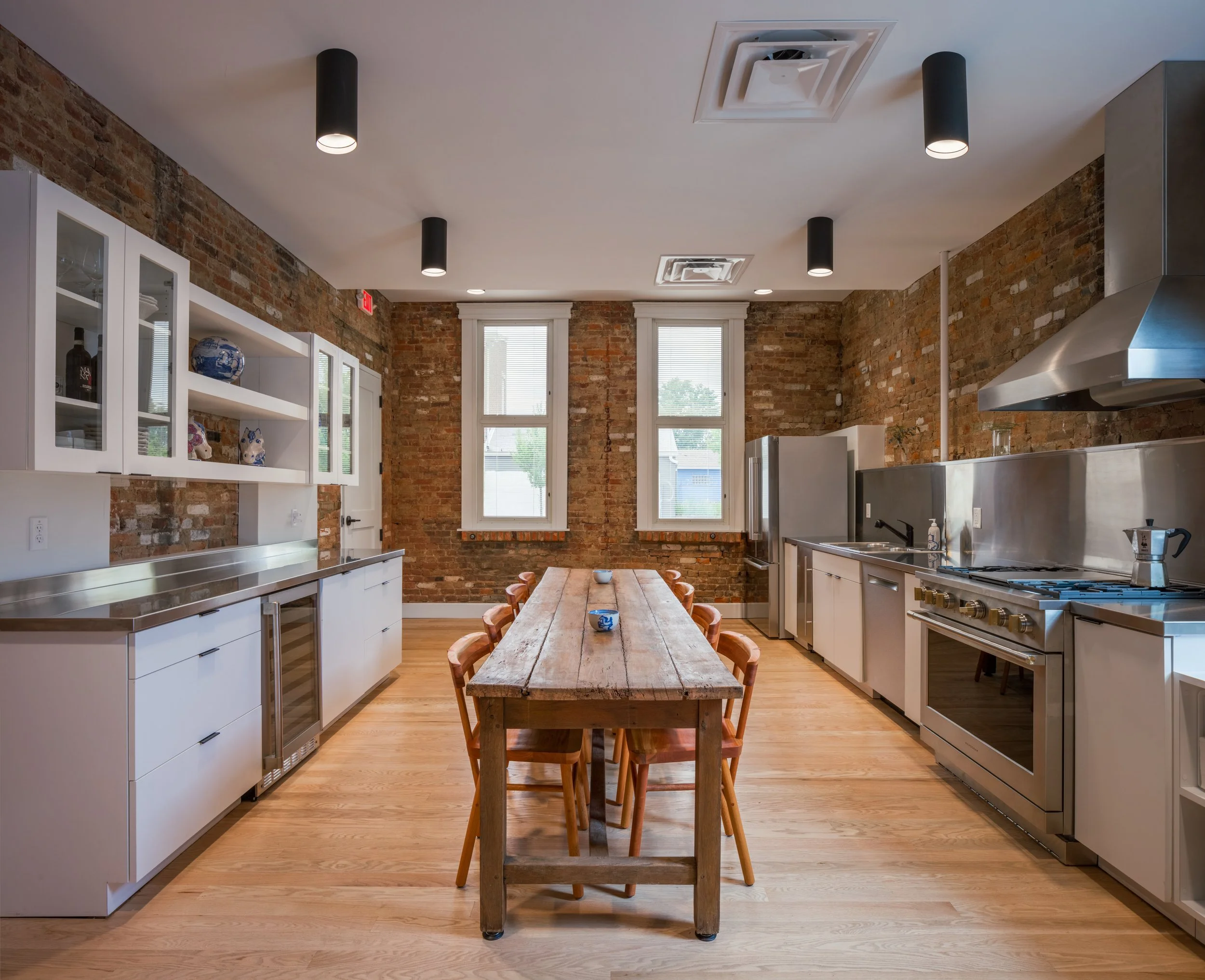 Modern kitchen with exposed brick walls, white cabinets, a wooden dining table, and black ceiling lights.