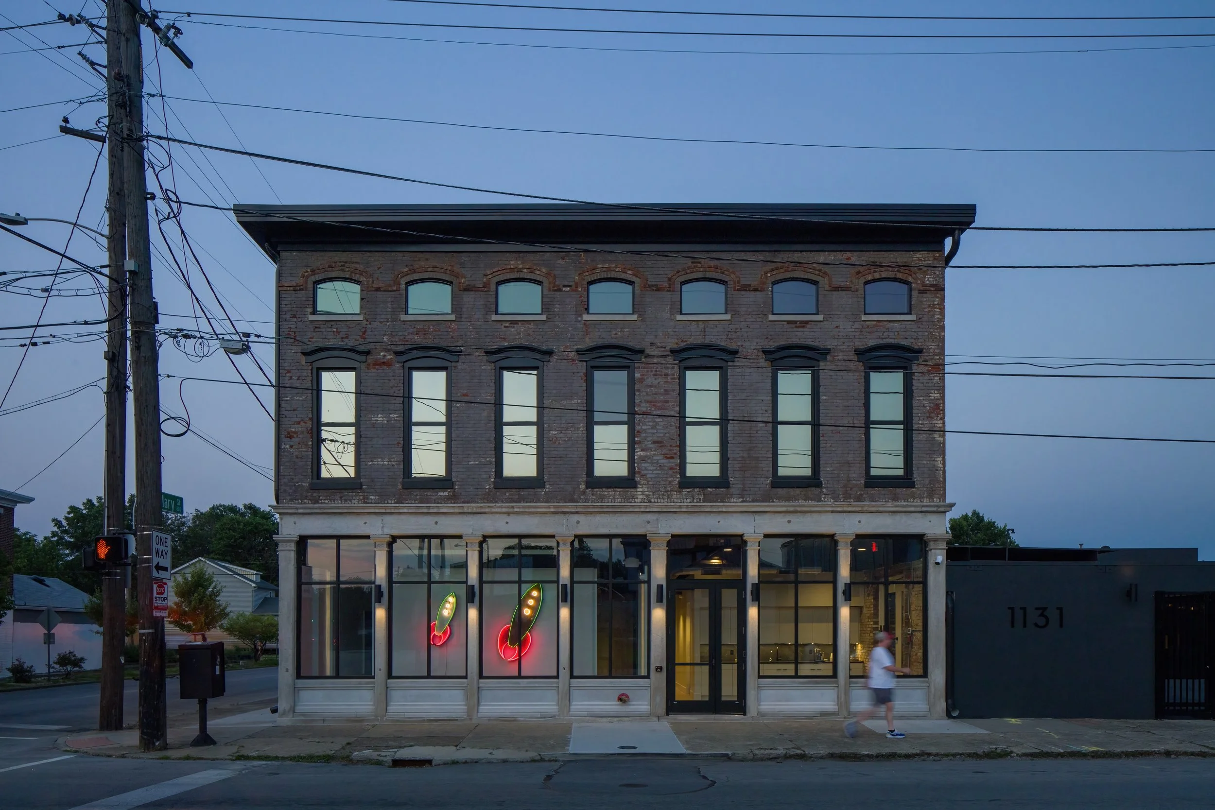 A three-story brick building with a storefront on the ground level, featuring large glass windows with neon cactus signs, and an apartment or office space on the upper floors, with multiple arched and rectangular windows; utility poles and wires in the foreground, a pedestrian crossing sign, and a traffic signal showing red hand signal, as well as a person walking on the sidewalk in front of the building, with trees and houses visible in the background, under a clear blue sky.