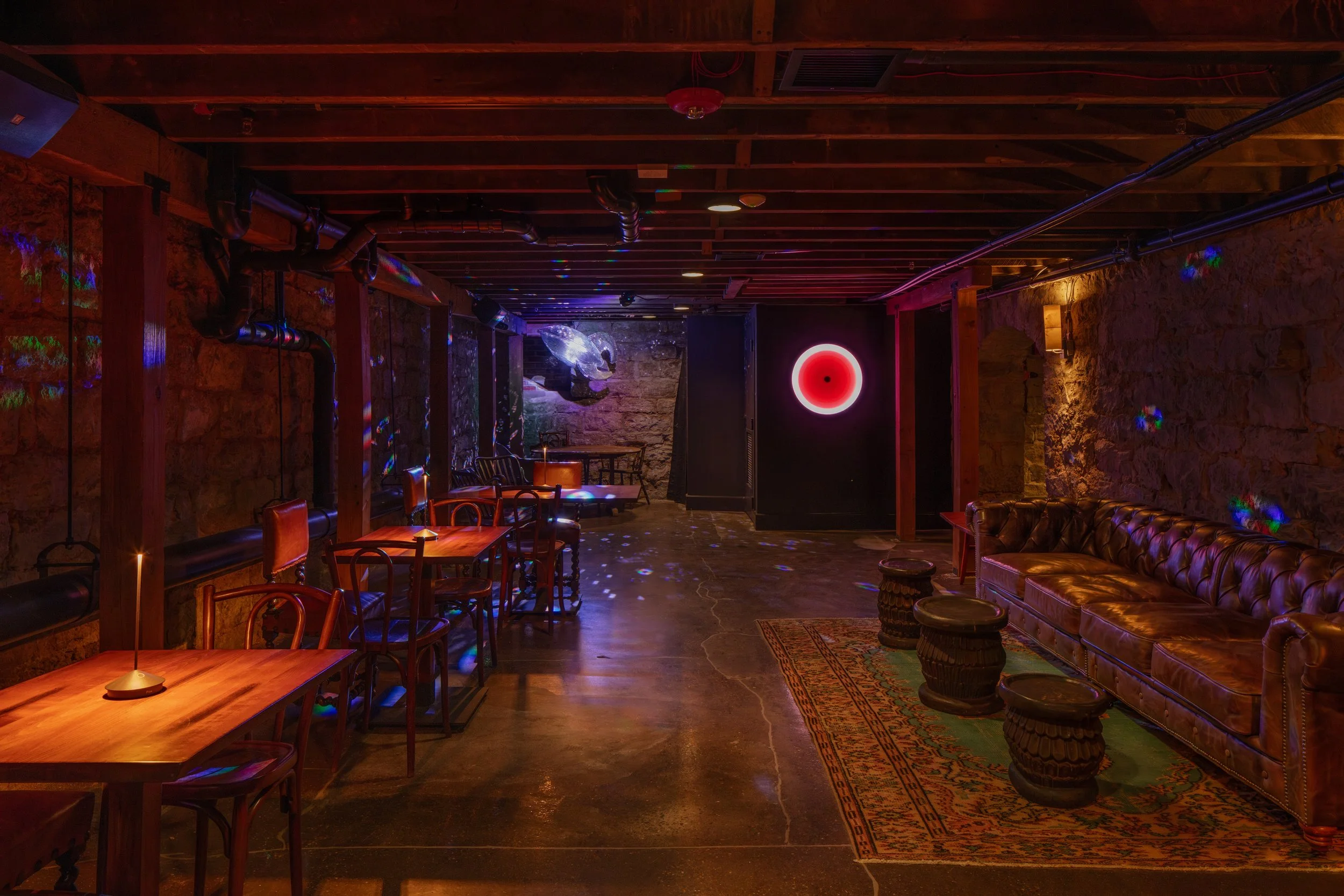 A dimly lit lounge area with wooden tables and chairs, exposed brick walls, a leather sofa, and colorful disco lights on the ceiling.
