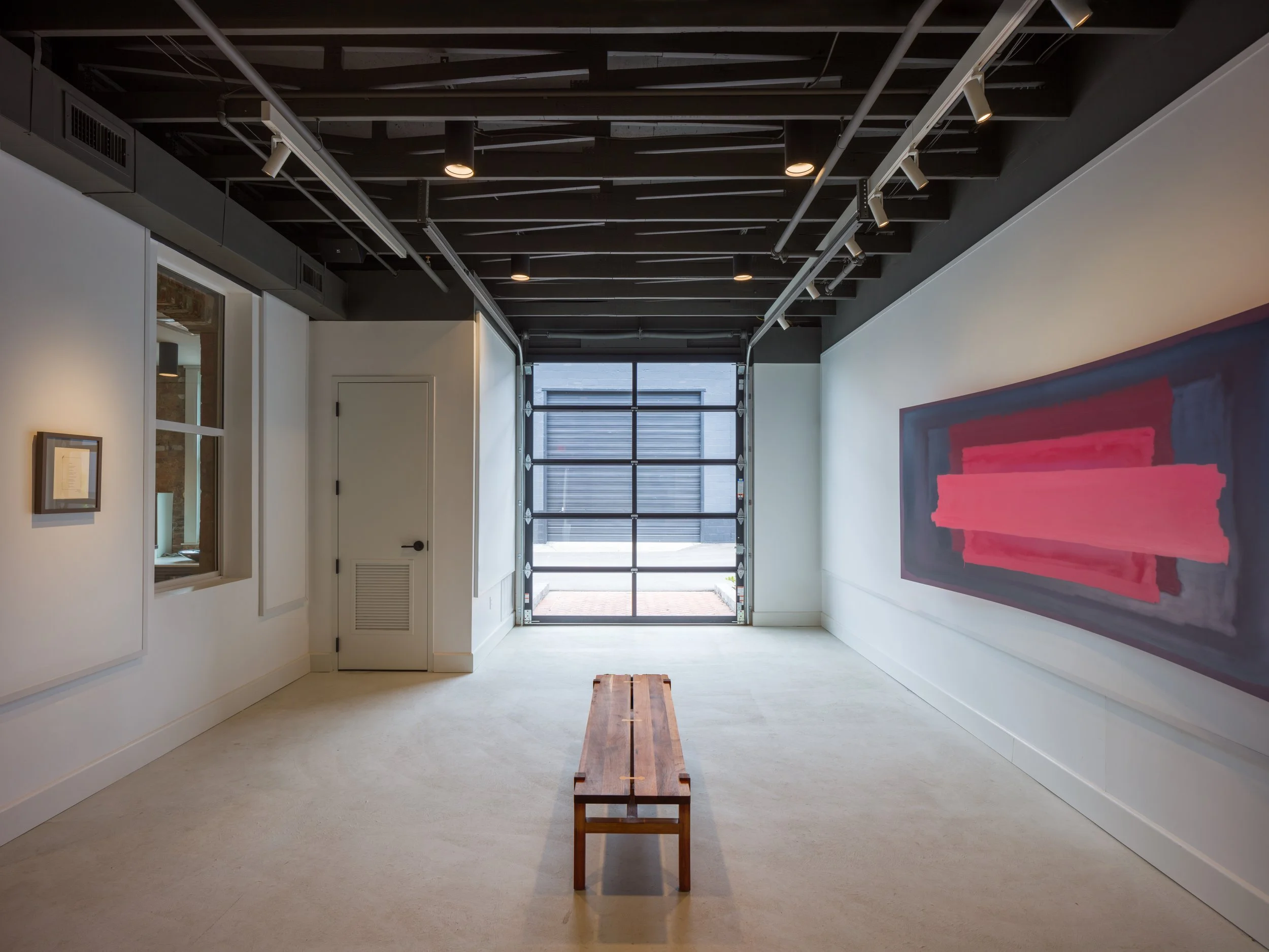 Empty gallery with white walls, black ceiling with exposed beams and track lighting, a large colorful abstract painting on the right, a wooden bench in the center, and a large window or garage door at the back.