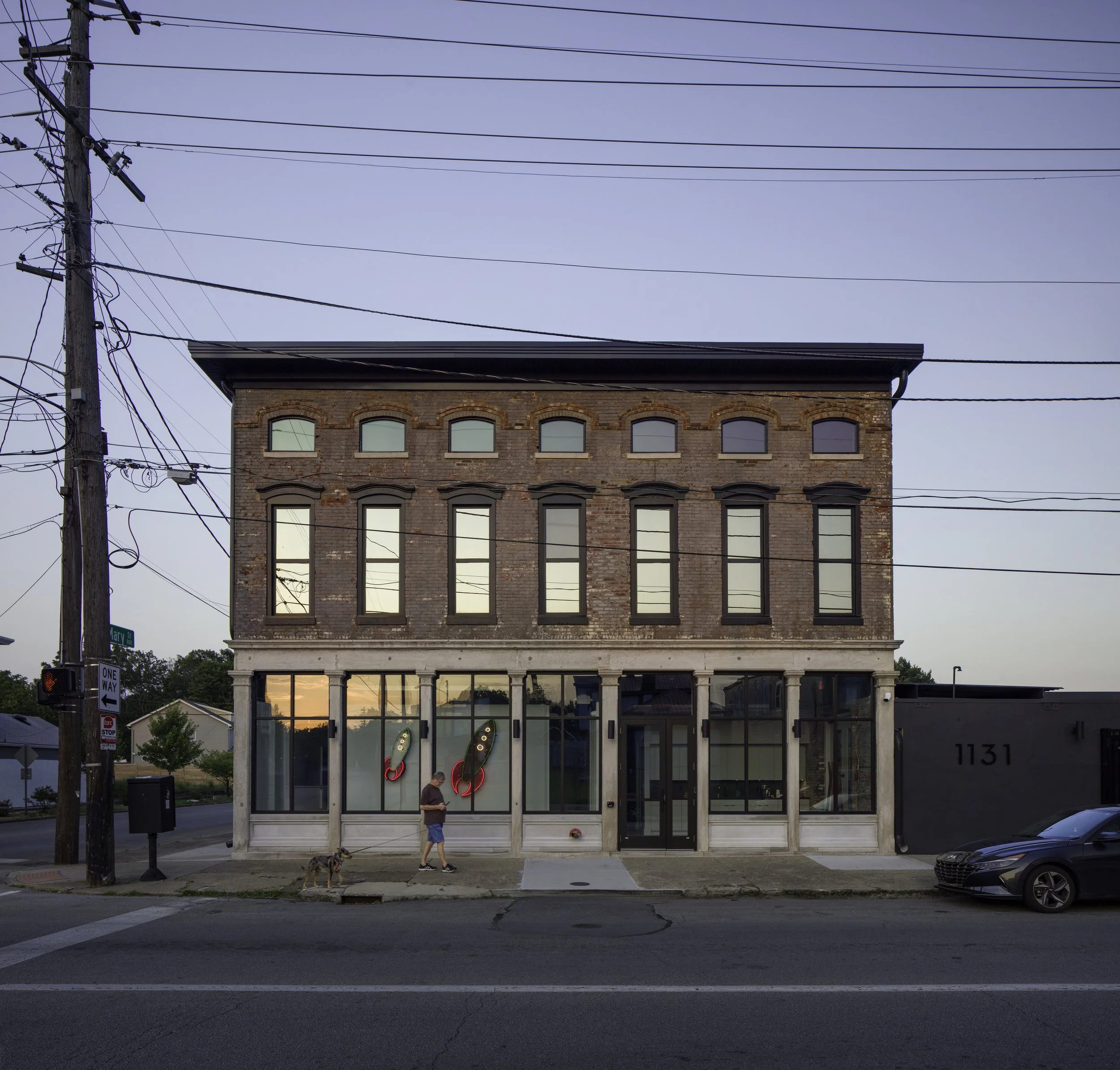 A three-story brick building with large glass windows on the first floor and smaller arched windows on the upper floors. A person is walking outside the building with a dog. Neon ice cream cone signs are visible inside the window. The scene is during dusk with a clear sky. A street corner with traffic lights, street signs, and parked cars is in the foreground.