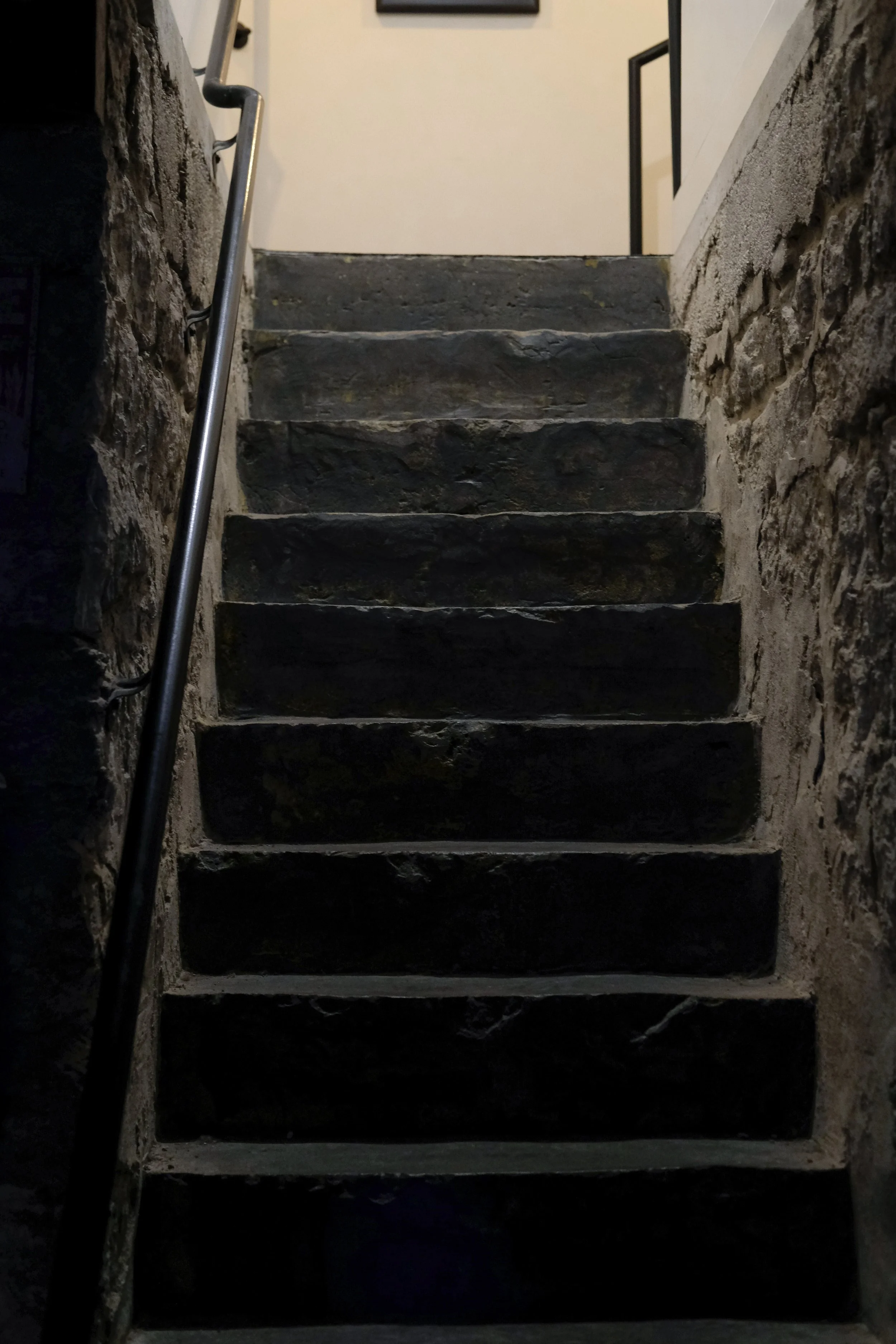 Stone staircase with metal handrail, leading up to a doorway with a white wall and black railing in the background.