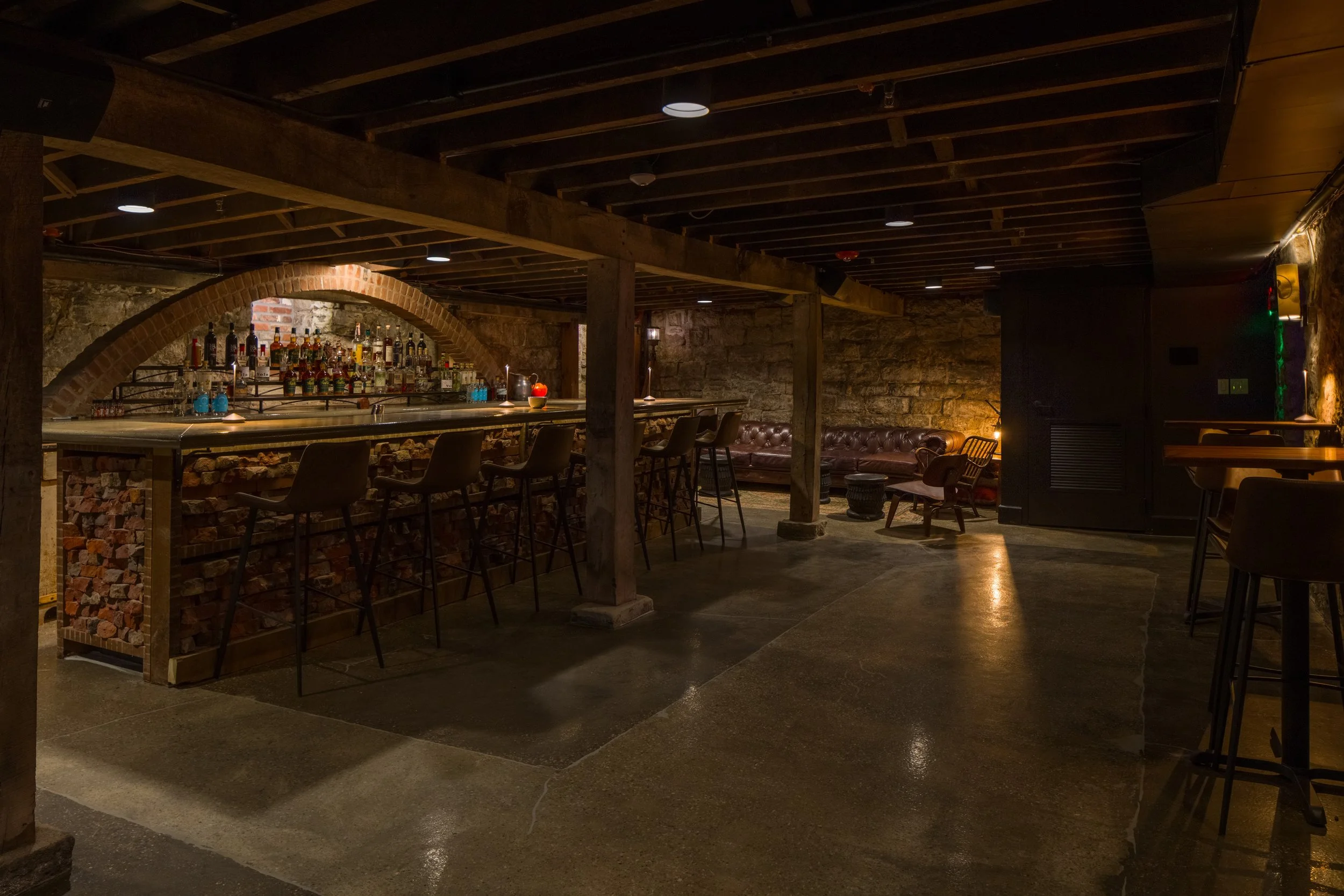 Dimly lit bar with exposed brick walls, wooden ceiling beams, a countertop with various bottles of liquor, and a cozy seating area with chairs and a sofa.