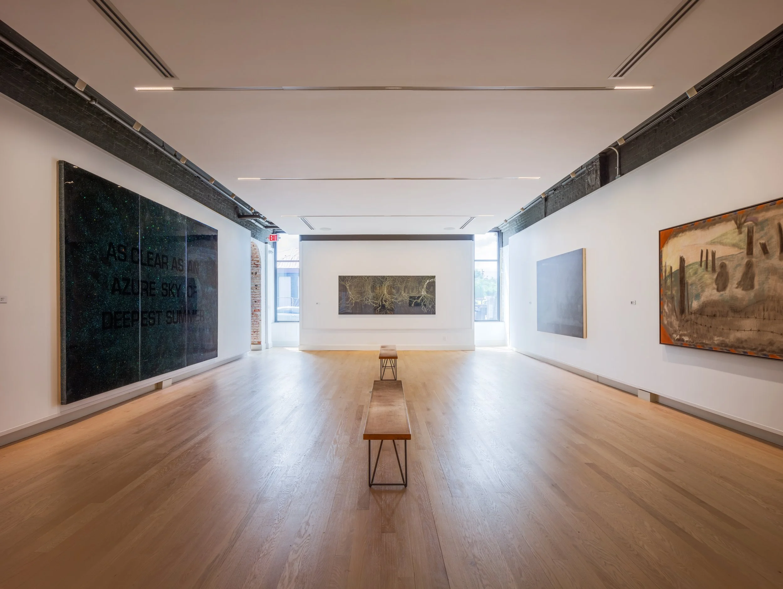 An art gallery with white walls and wooden floors, featuring large paintings on the walls, a small wooden bench in the center, and large windows at the back letting in natural light.