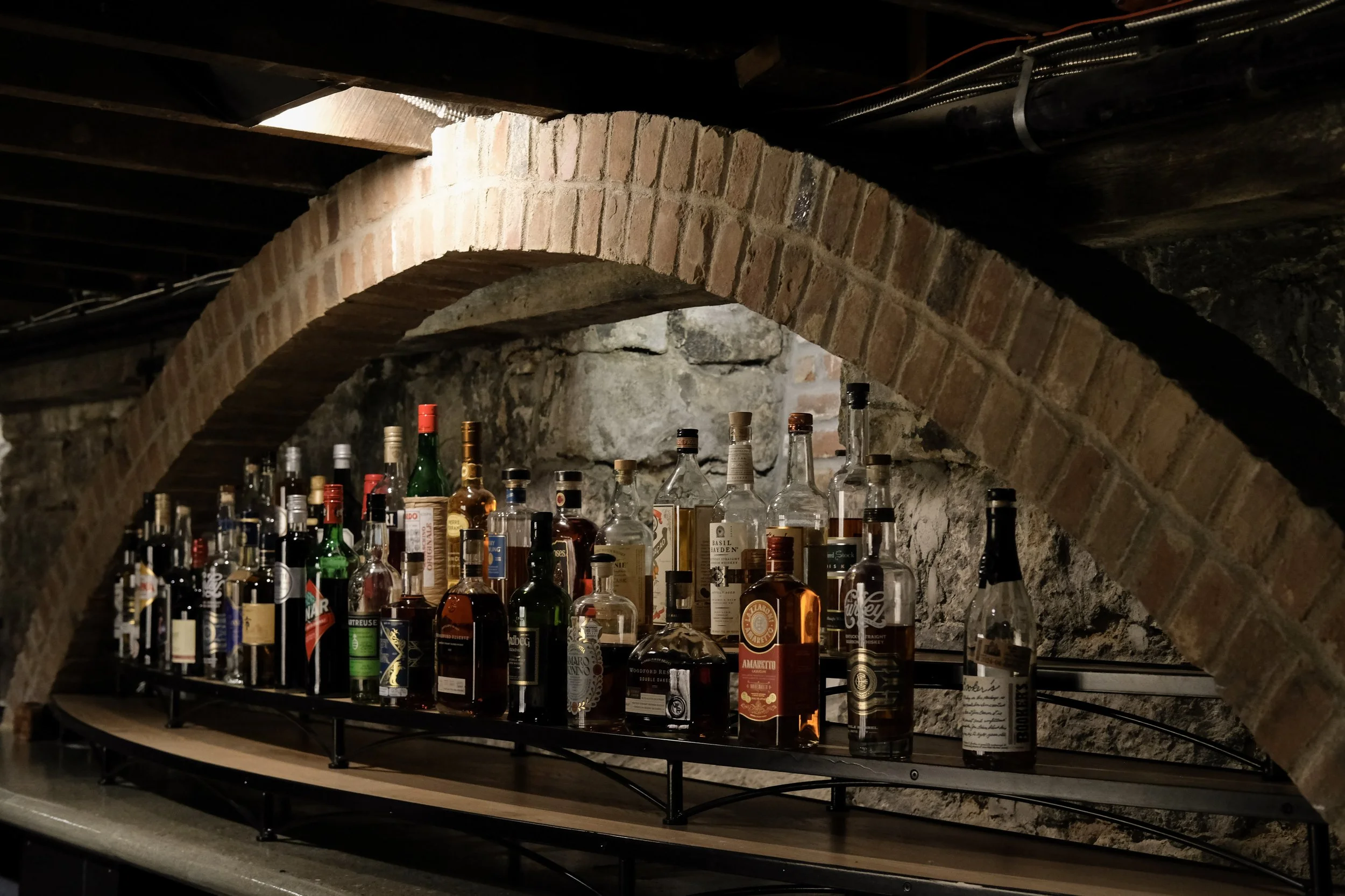 A collection of assorted liquor bottles on a bar shelf beneath an arched brick ceiling in a basement or cellar setting.