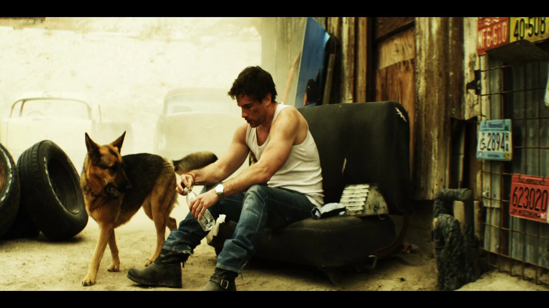Man in a white tank top sitting on a black couch in a rustic garage setting with a German Shepherd dog, surrounded by old tires and vintage license plates.