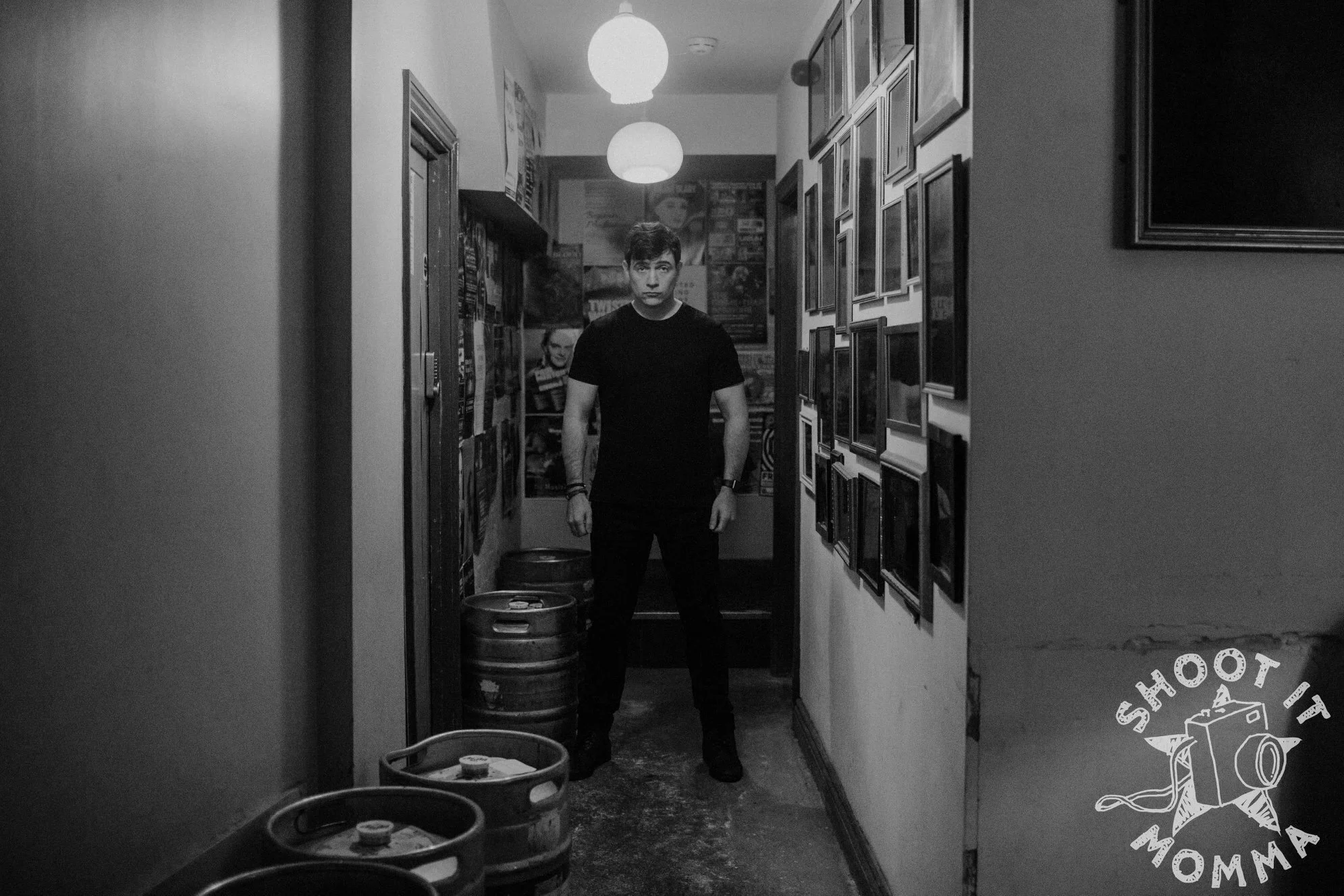 Black and white photo of a man standing in a narrow hallway with kegs and framed pictures on the walls.