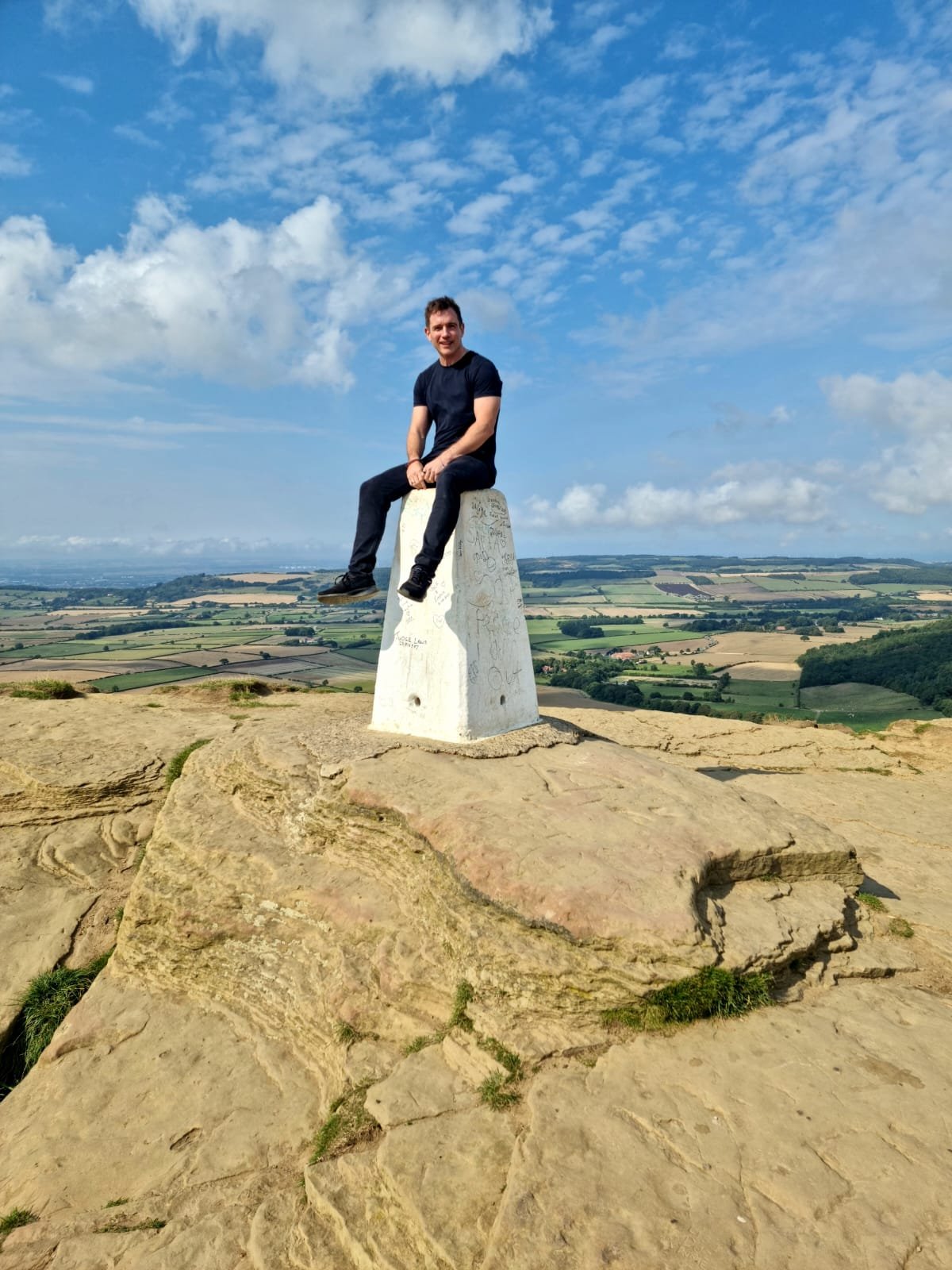 Person sitting on a trig point on a rocky hilltop with a panoramic rural landscape in the background under a blue sky with scattered clouds.
