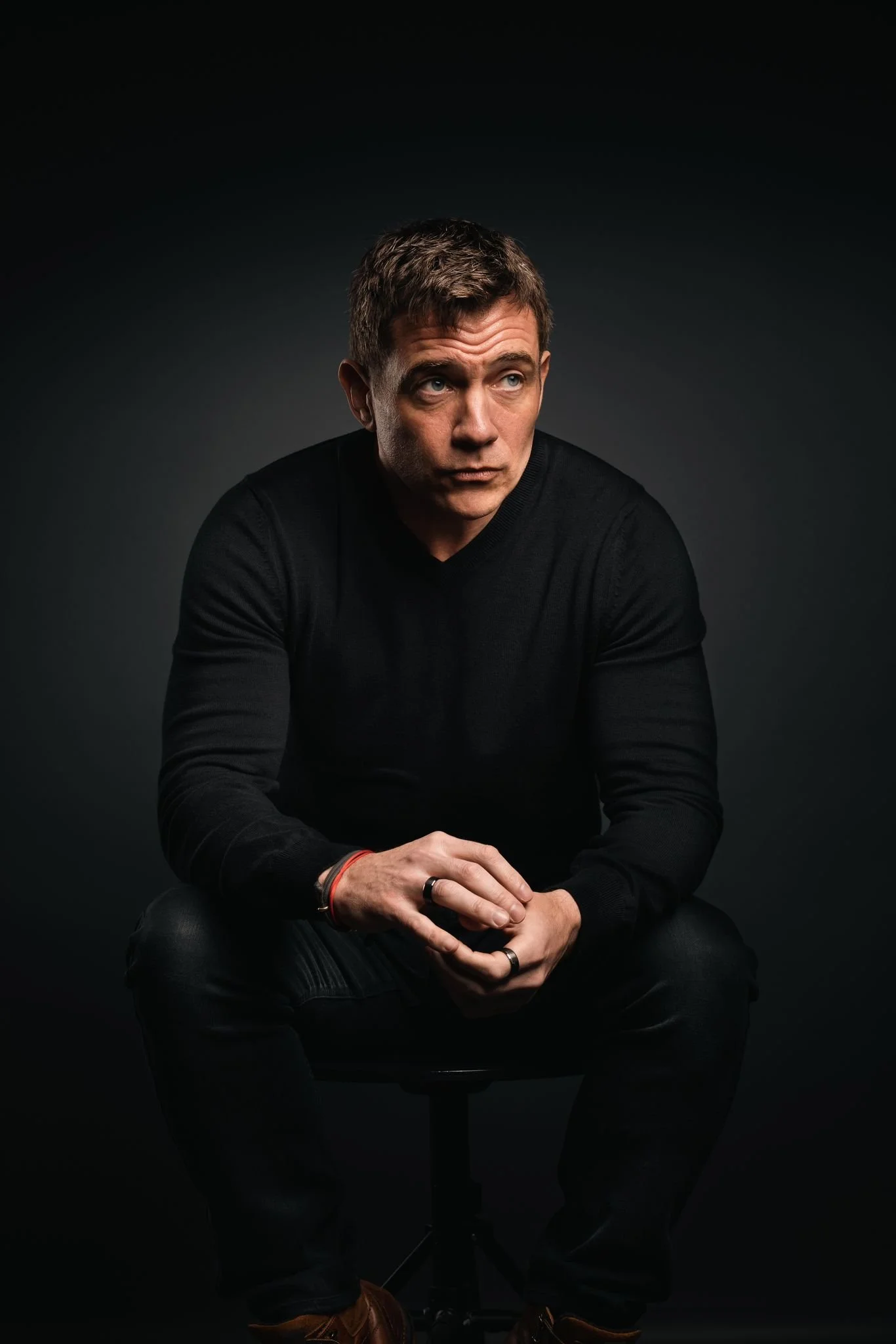 Paul cashmore young man with short brown hair, wearing a black sweater, sits on a stool against a dark background. He has a thoughtful expression, with his hands clasped together, adorned with rings and bracelets.