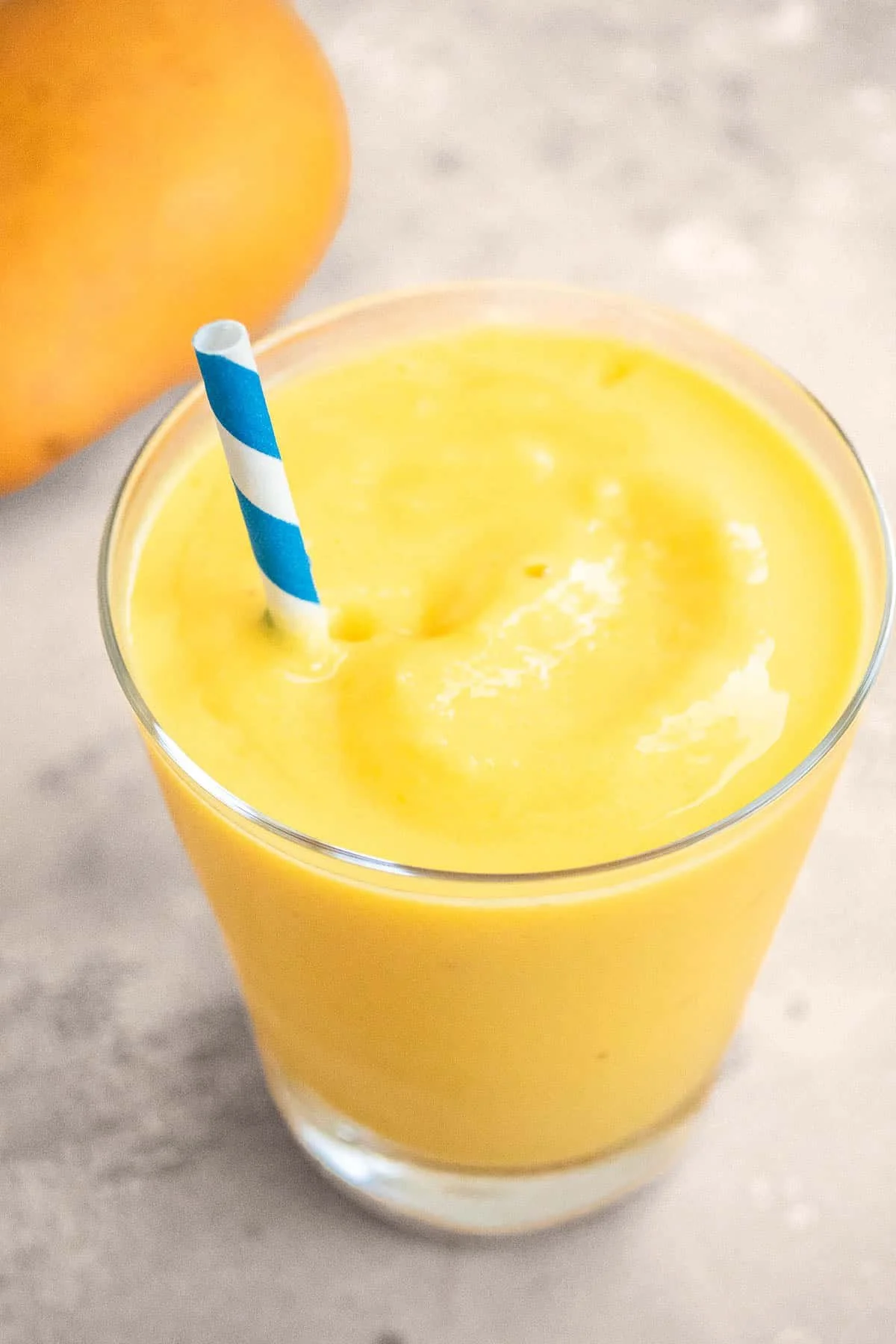 Mango smoothie in a glass with a blue and white striped straw on a concrete surface.