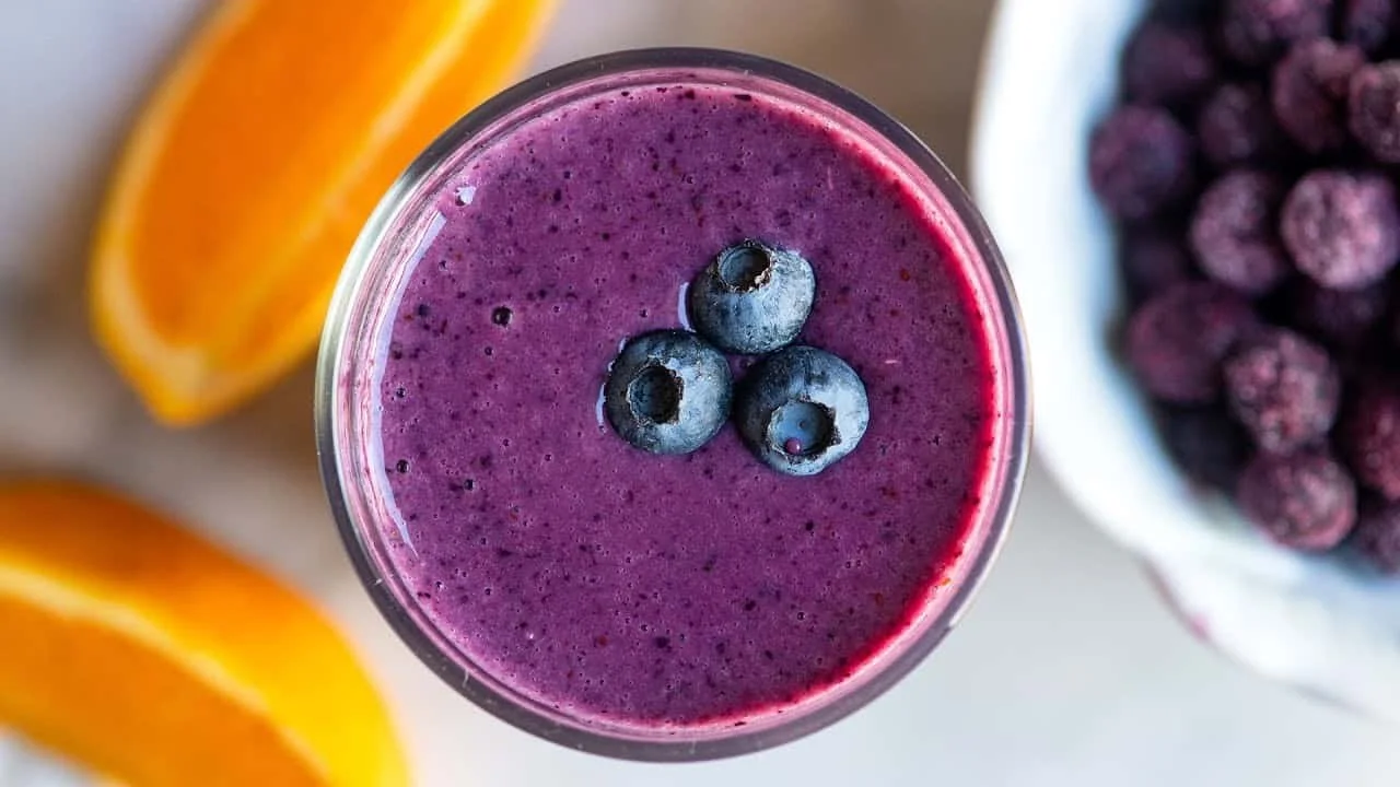 A glass of purple blueberry smoothie topped with three blueberries, with orange slices and a bowl of frozen berries in the background.