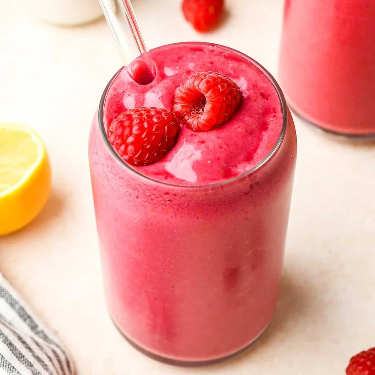 Glass filled with pink raspberry smoothie topped with fresh raspberries and a glass straw, surrounded by lemon half and cloth napkin on a light surface.