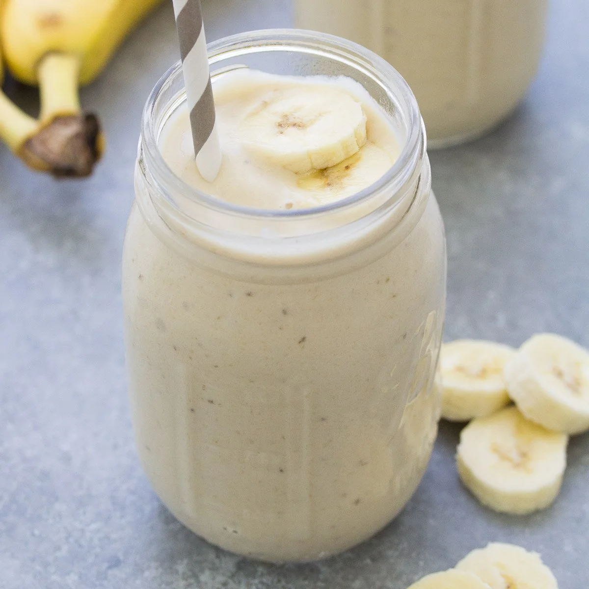 Banana smoothie in a glass jar with sliced bananas and a striped straw on a gray surface.