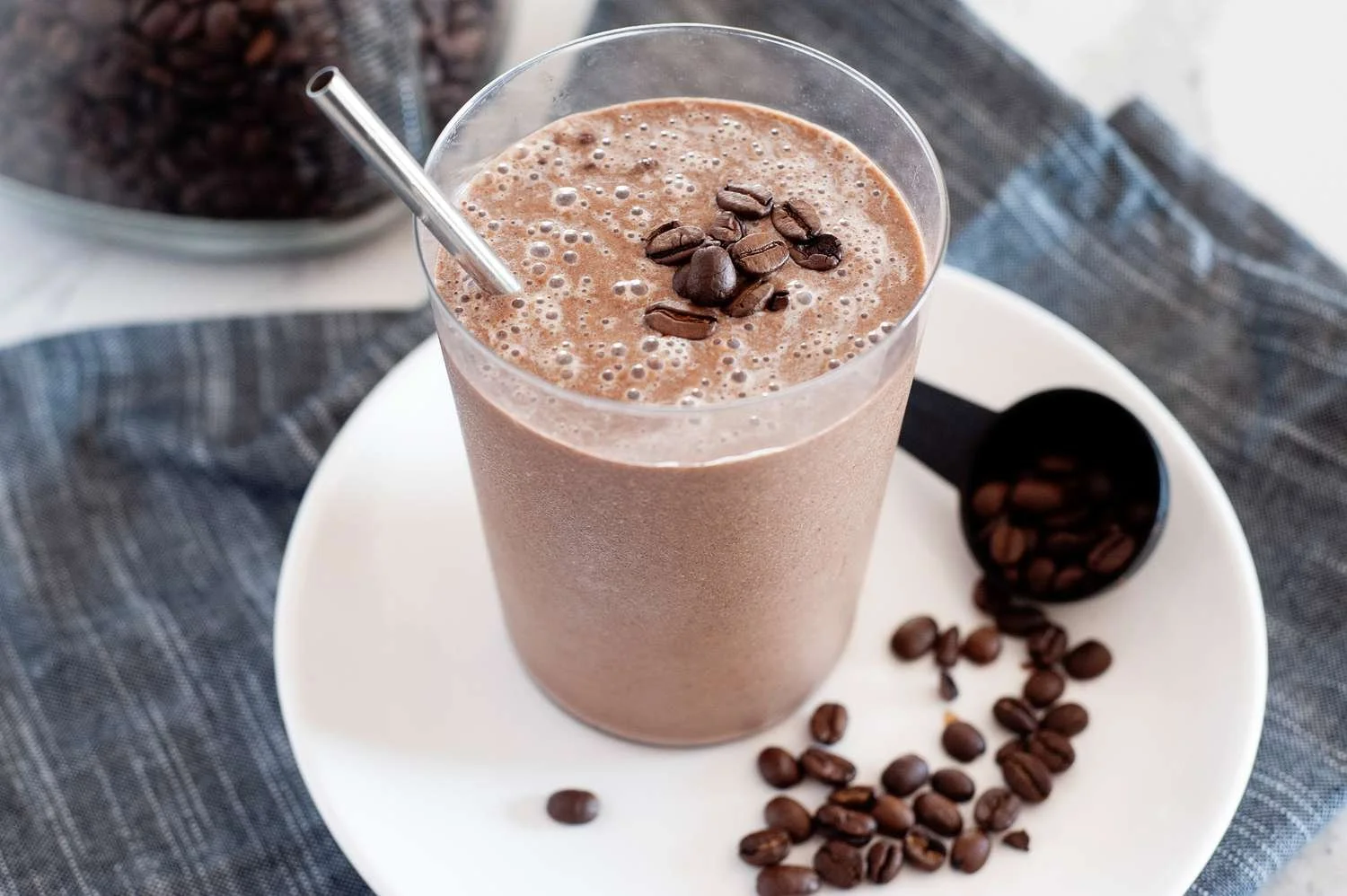 Iced coffee smoothie in a glass with a metal straw, topped with coffee beans, on a white plate with scattered coffee beans and a scoop, placed on a blue fabric.