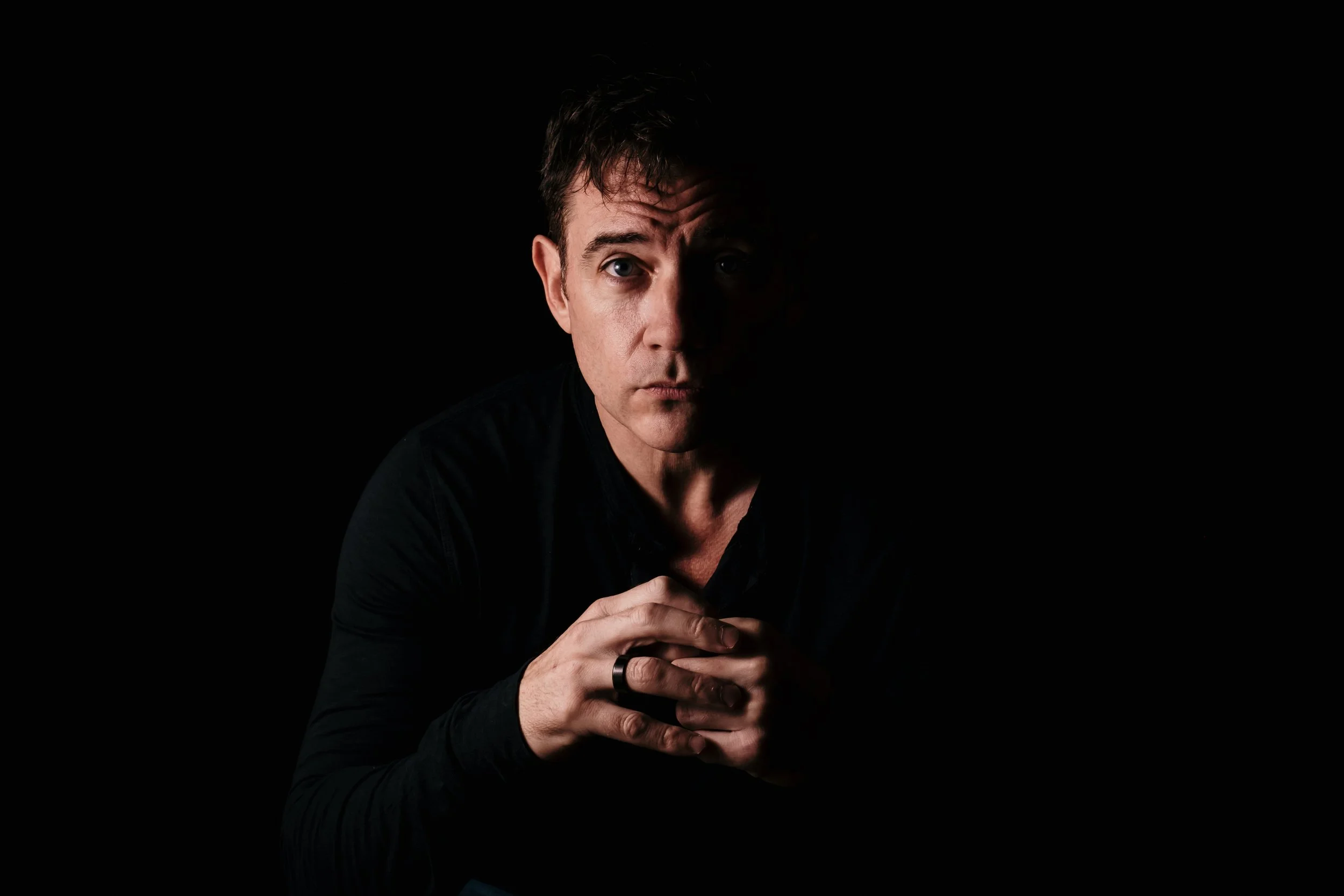 Man in dark lighting with hands clasped and serious expression