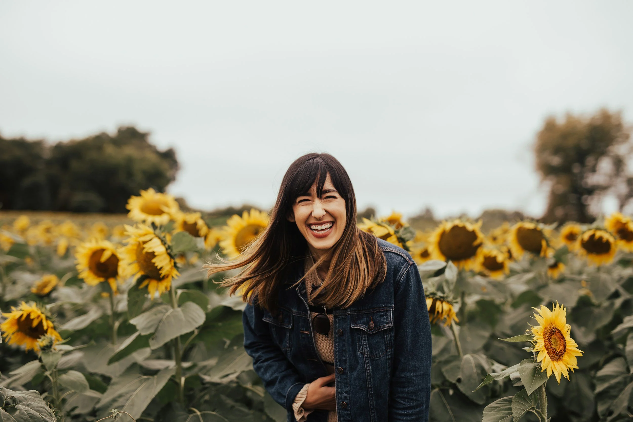 A woman with long brown hair, smiling broadly, wearing a denim jacket, standing in a sunflower field with trees in the background on a cloudy day.