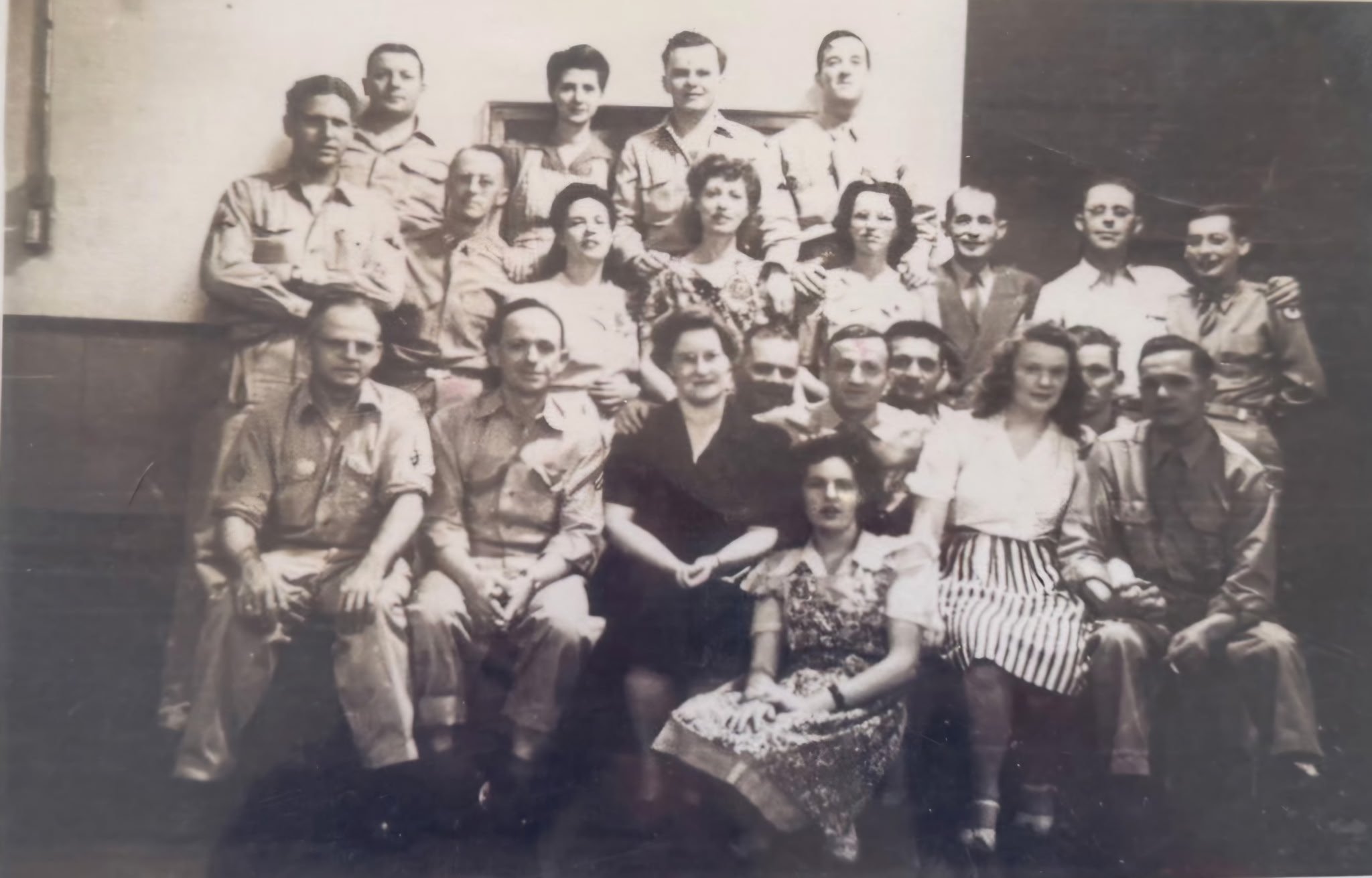 Black and white historical photograph of 16 people, including men and women, posing outdoors in front of a building. Some individuals are wearing military uniforms, while others are in casual or civilian clothing. The group includes a woman holding a floral bouquet and a young girl in a striped skirt. The scene appears to be a commemorative or social gathering.