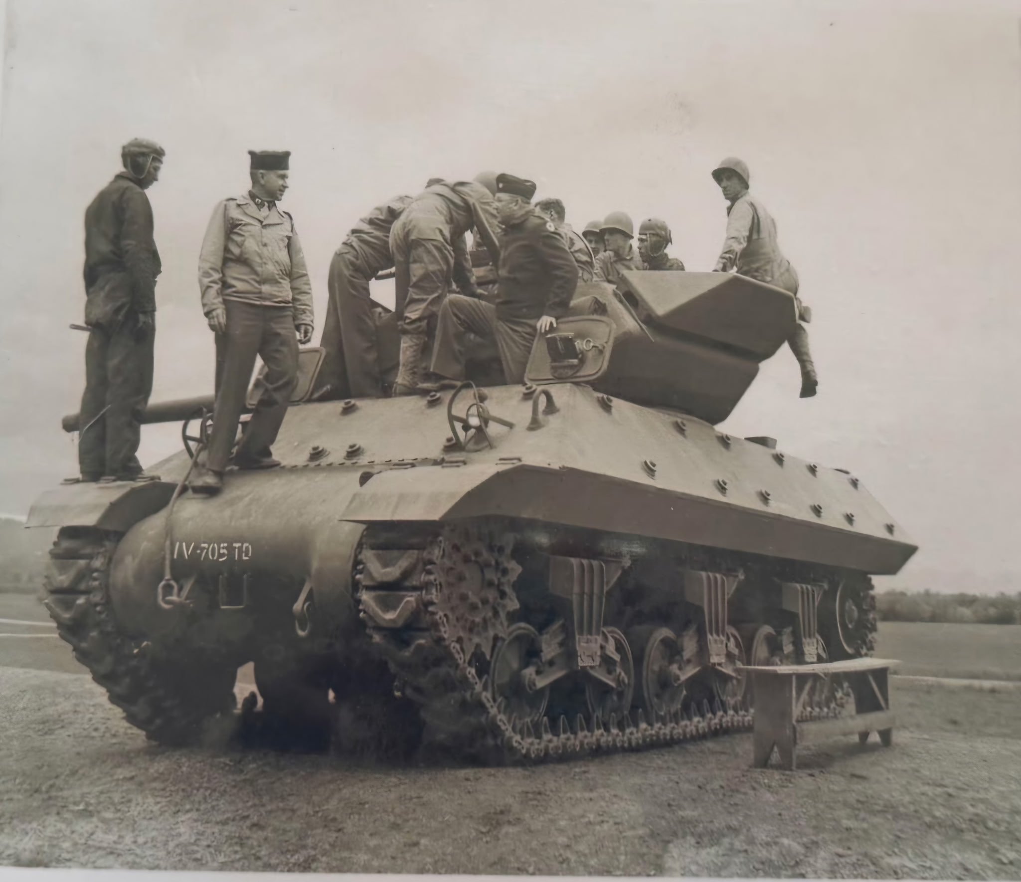 A group of soldiers gathered around an armored tank, inspecting or preparing it, with some soldiers on top and others standing nearby, in an outdoor setting.