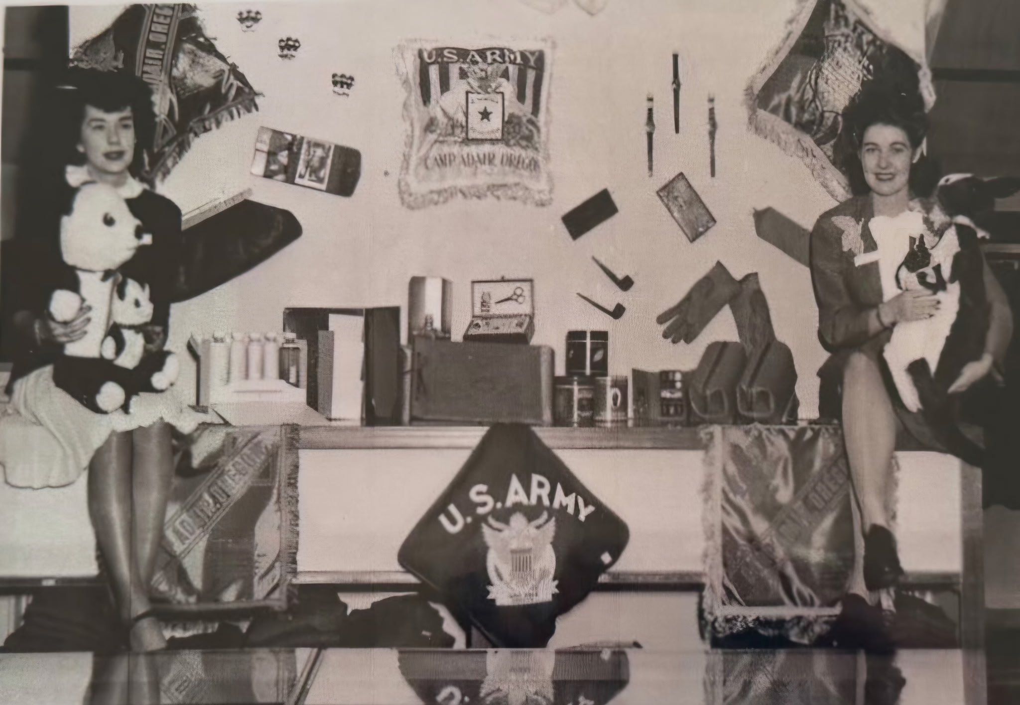 Black and white photograph of two women in vintage clothing posing in front of a U.S. Army display with flags, a badge, and various military items.