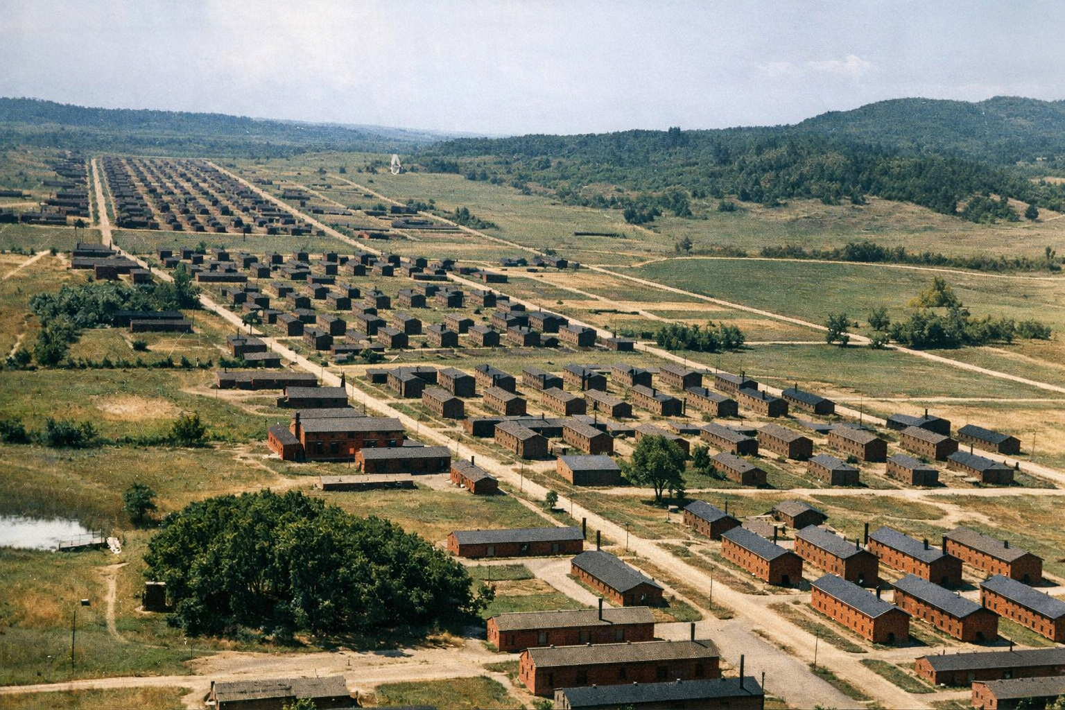 Aerial view of multiple rows of brick buildings in a sprawling camp or settlement, surrounded by greenery and hills in the background.