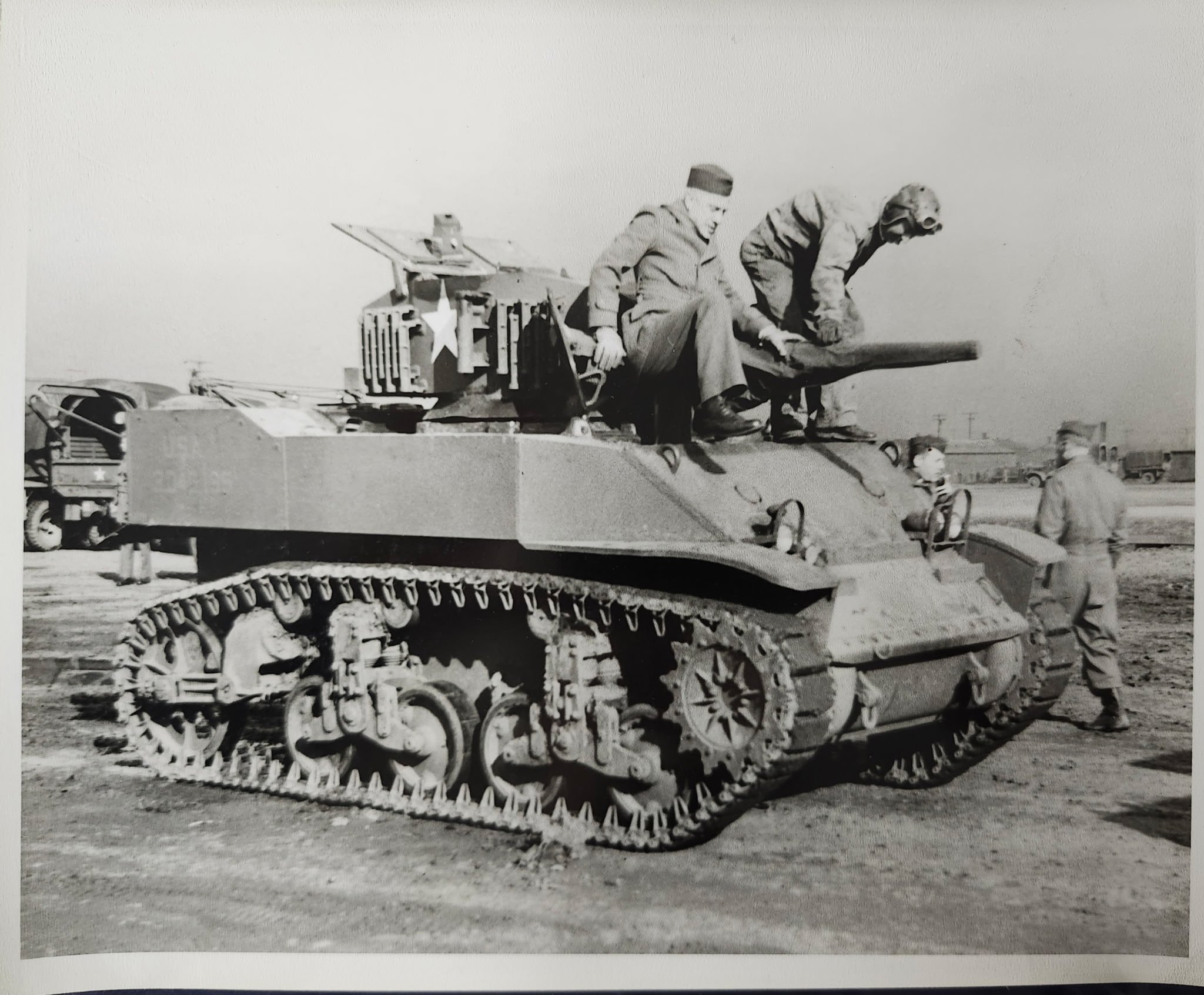 Black and white photograph of a tank with two soldiers sitting on top, one in a military uniform and the other wearing a hood. The tank's turret features a star emblem, and the background includes vehicles and a flat landscape.