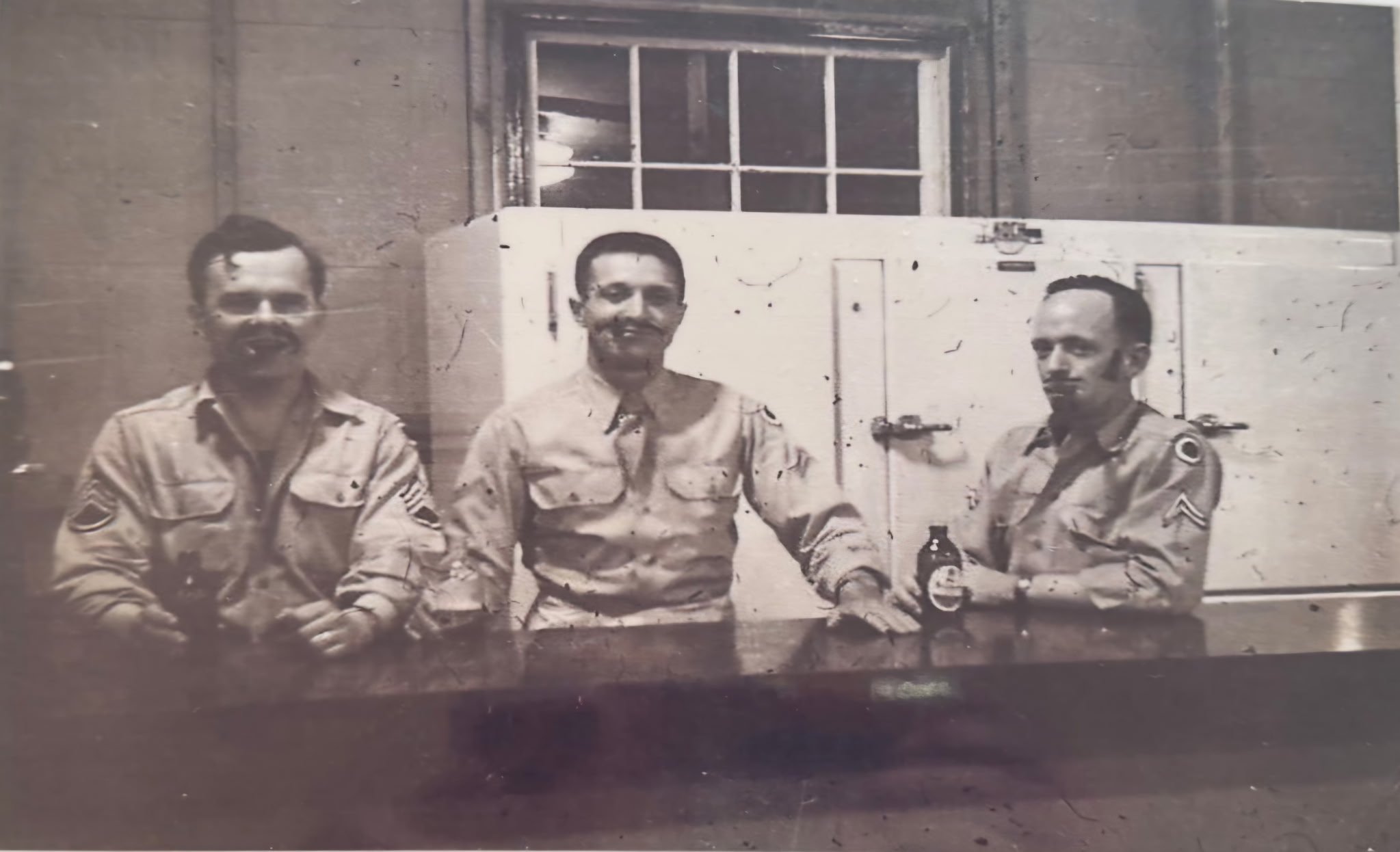 Three men in military uniforms sitting at a table with a bottle and glasses, in front of a window and wood-paneled wall.