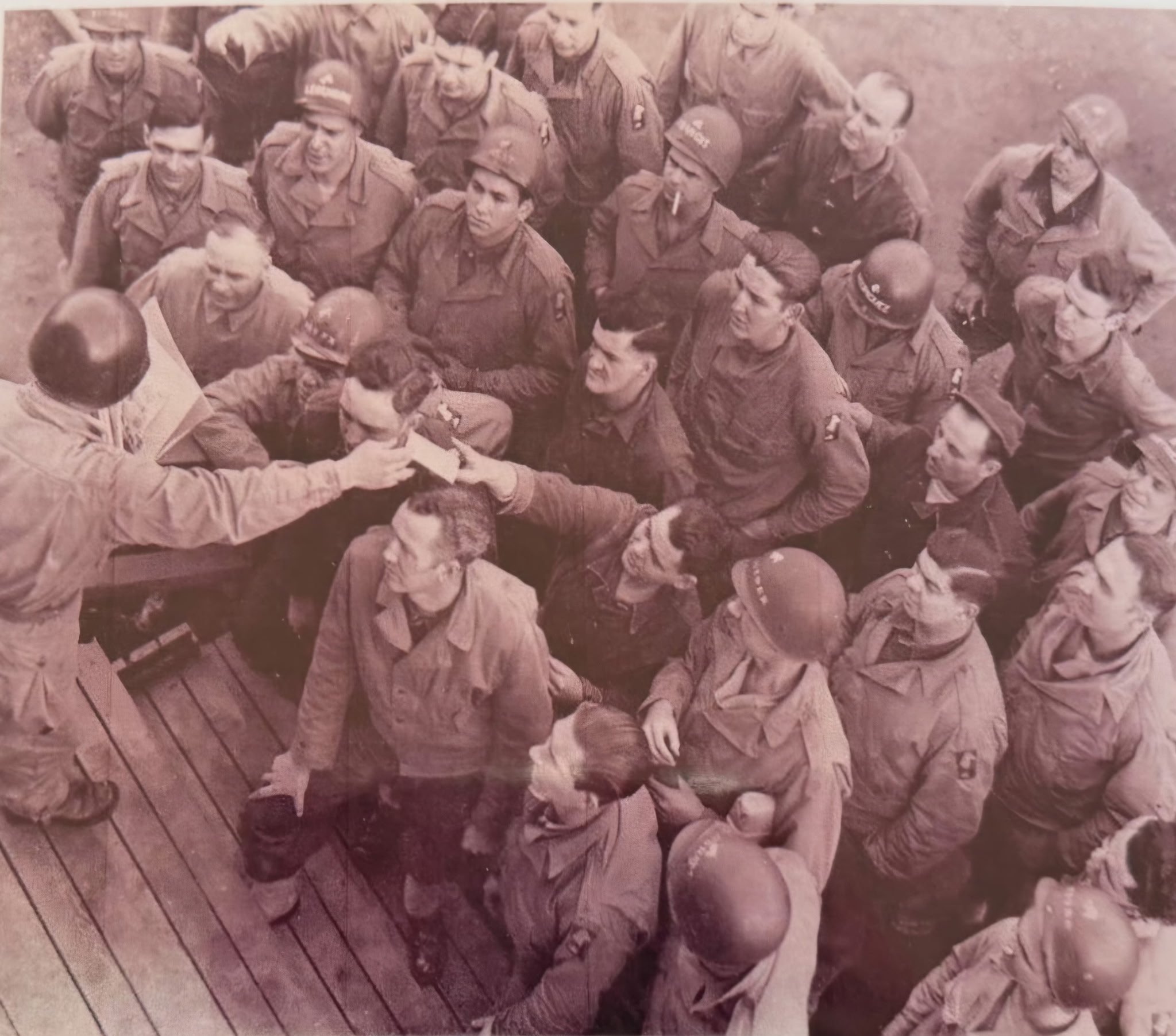 Black-and-white photograph of a group of soldiers gathered outdoors, some sitting and some standing, with a wooden deck in the foreground.