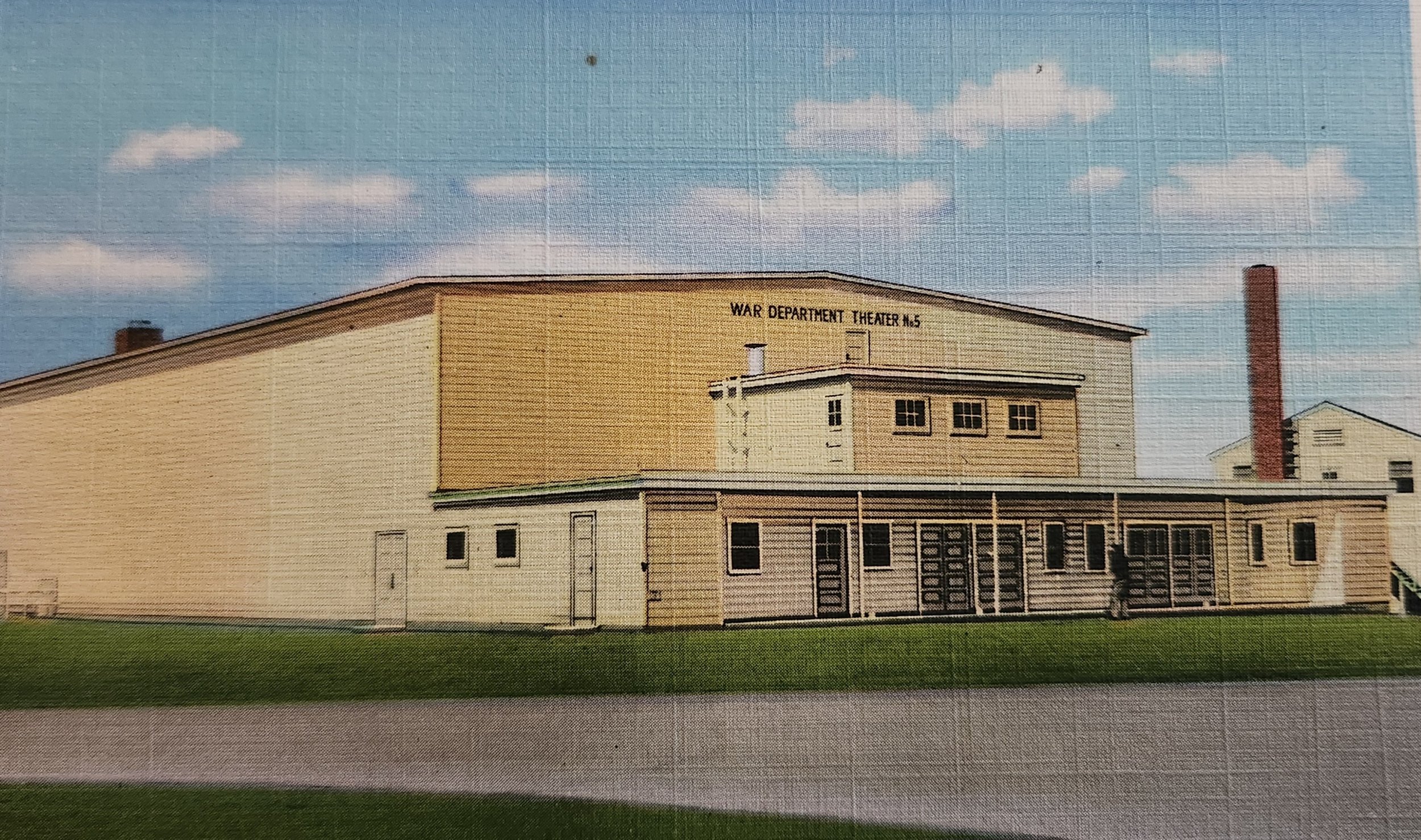 A large beige building with a sign that reads 'War Department Theater No. 5' on the top right. The building has a sloped roof and multiple windows. There is a chimney and a small structure attached to the building. The sky above is partly cloudy with some in the background.