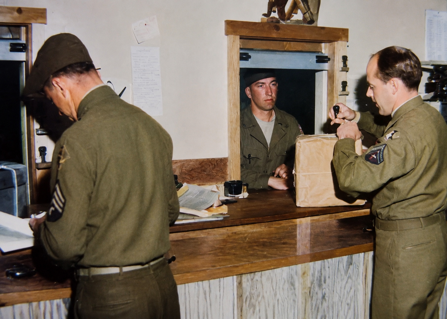 Three military personnel at a reception desk, with one man handling a package and another reading a document.