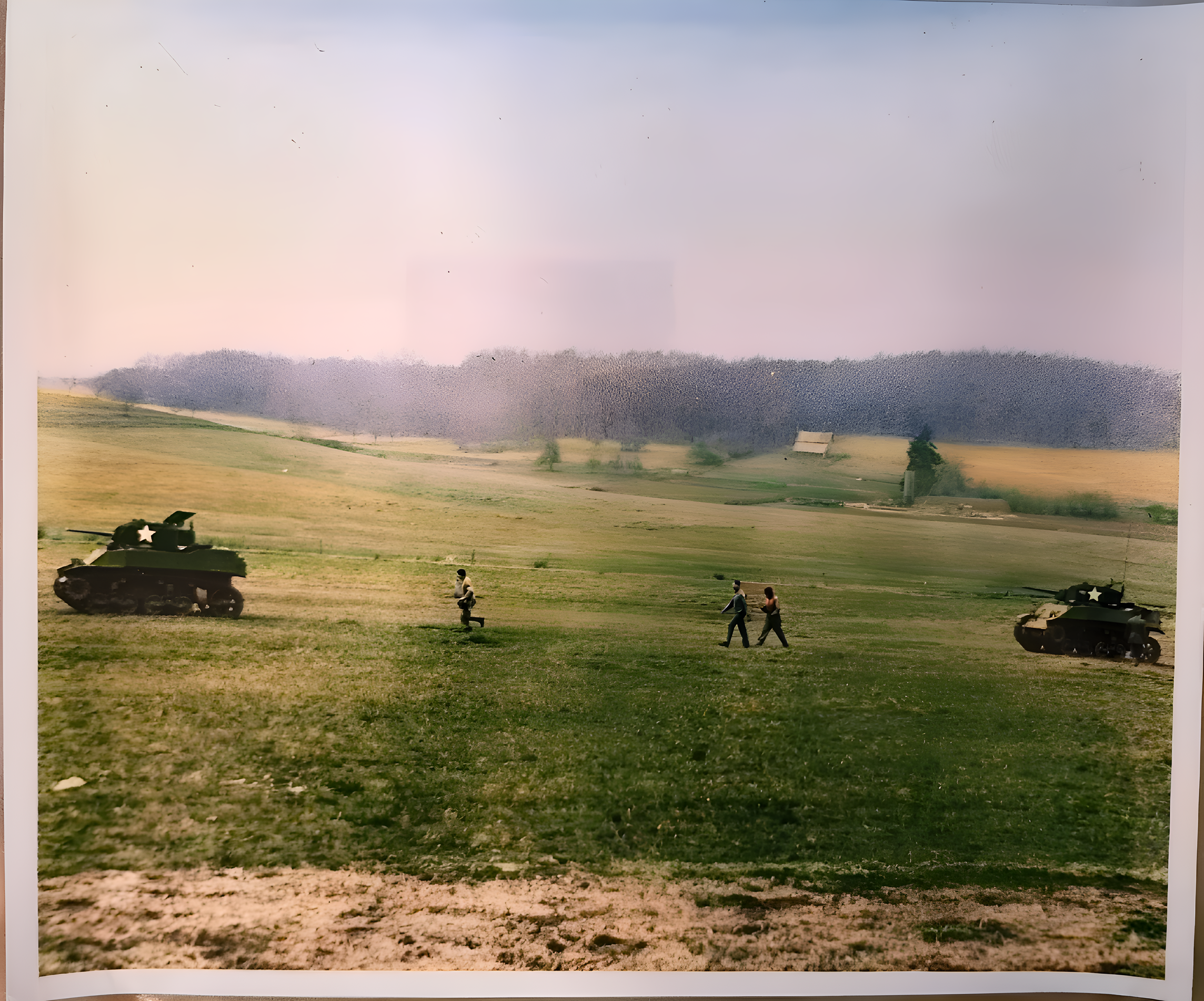 A landscape with a fence, trees, and a cloudy sky. Two military tanks are positioned on the field, with a helicopter flying in the sky.