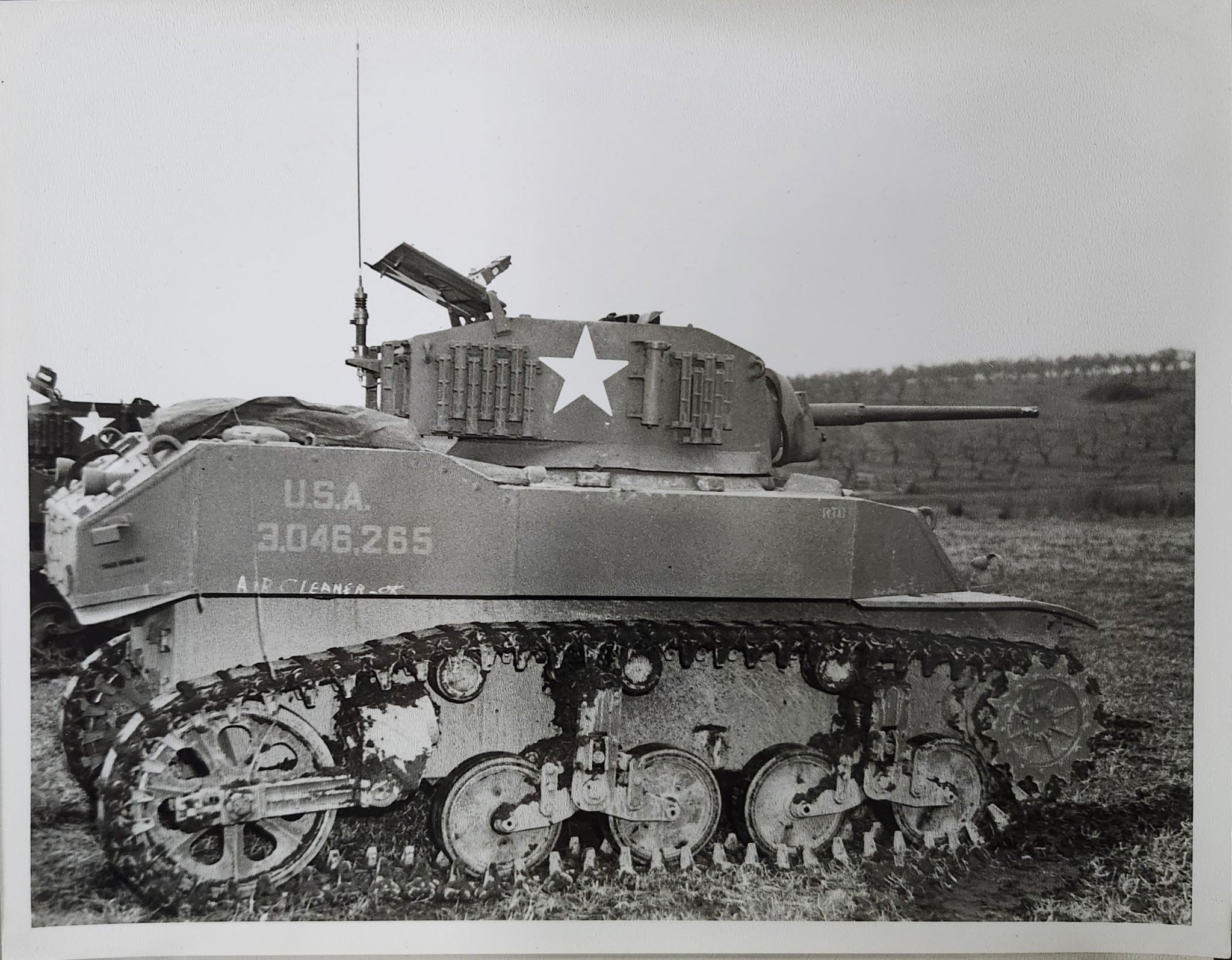 Vintage black and white photograph of a military tank with a large white star on its side and the markings "U.S.A." and "3046265" on its front, parked outdoors on a grassy field with a cloudy sky in the background.