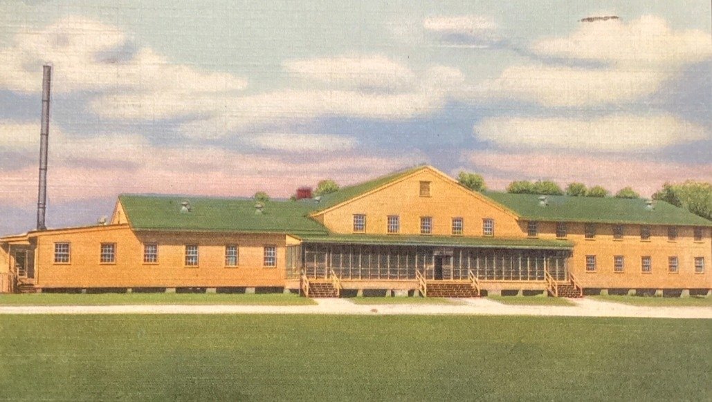 A large, yellow, single-story building with a green roof, multiple windows, and a screened porch in the center, with stairs leading up to it, set against a partly cloudy sky and a grassy lawn in the foreground.