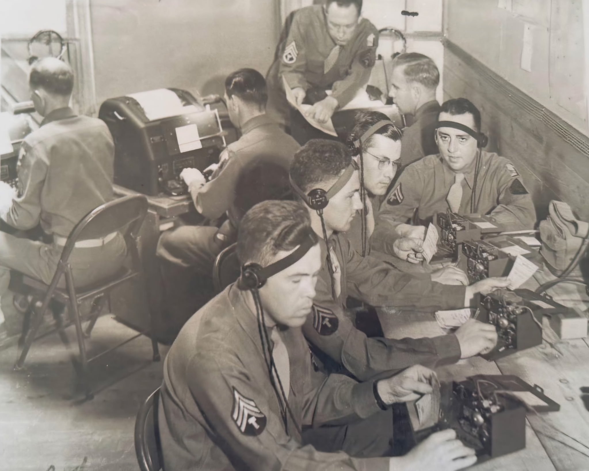 Black and white historical photograph of soldiers in uniform sitting and working around radios, with one soldier standing and handing a paper to another.