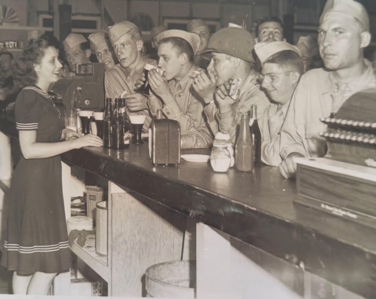 A young woman in a navy uniform working at a bar or soda fountain, serving a group of soldiers in military uniforms who are seated at the bar, some drinking and some ordering drinks, in what appears to be a busy, lively environment, possibly a restaurant or bar during wartime.