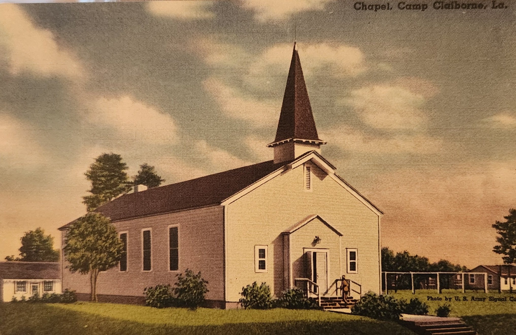 A church with a tall steeple, surrounded by trees and shrubs, with stairs leading to the entrance, and siding painted in pastel yellow.
