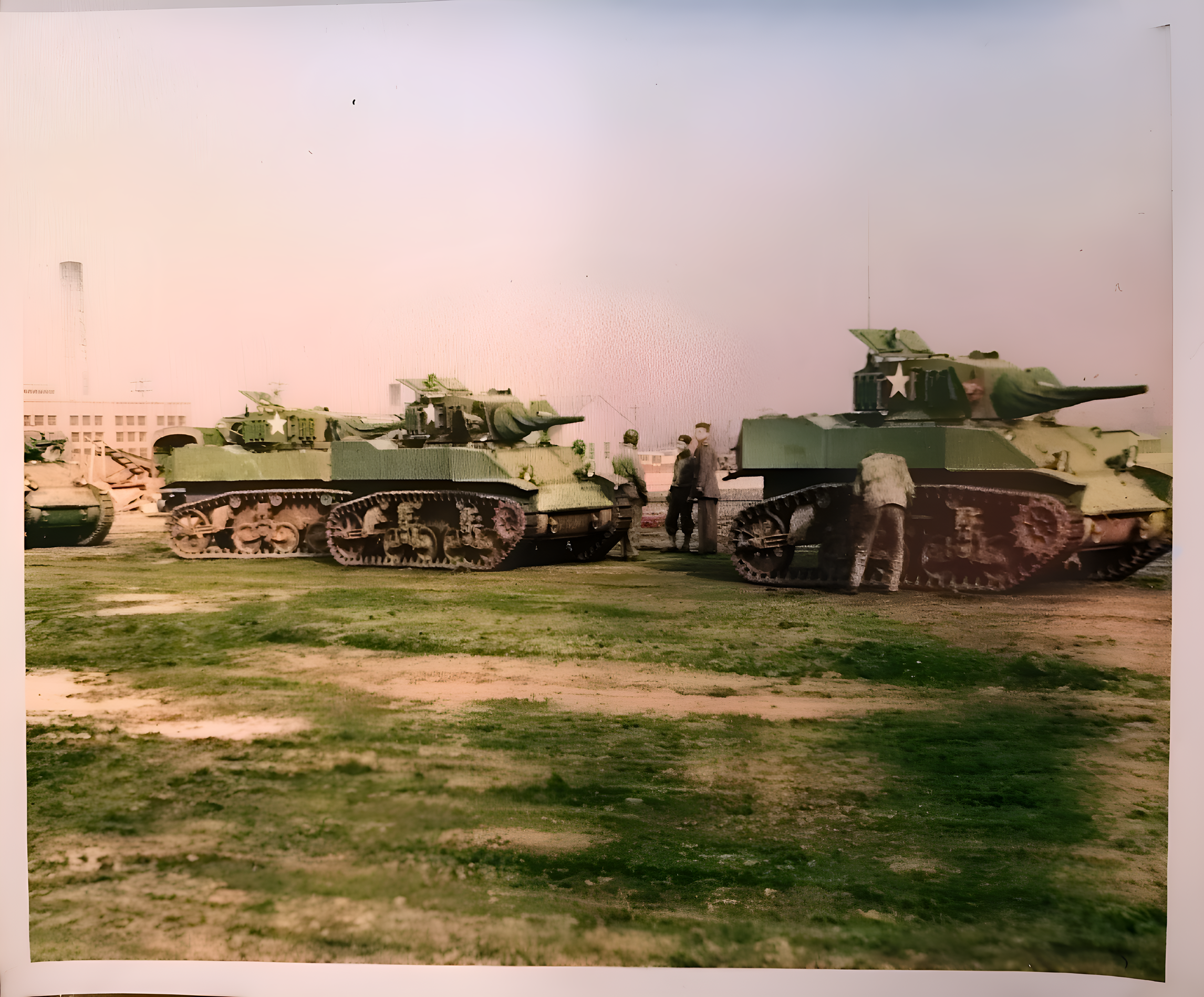 Several military tanks with green camouflage and white stars on the sides are lined up outdoors on a grassy area. There are a few people standing and walking near the tanks. Urban buildings are visible in the background under a cloudy sky.