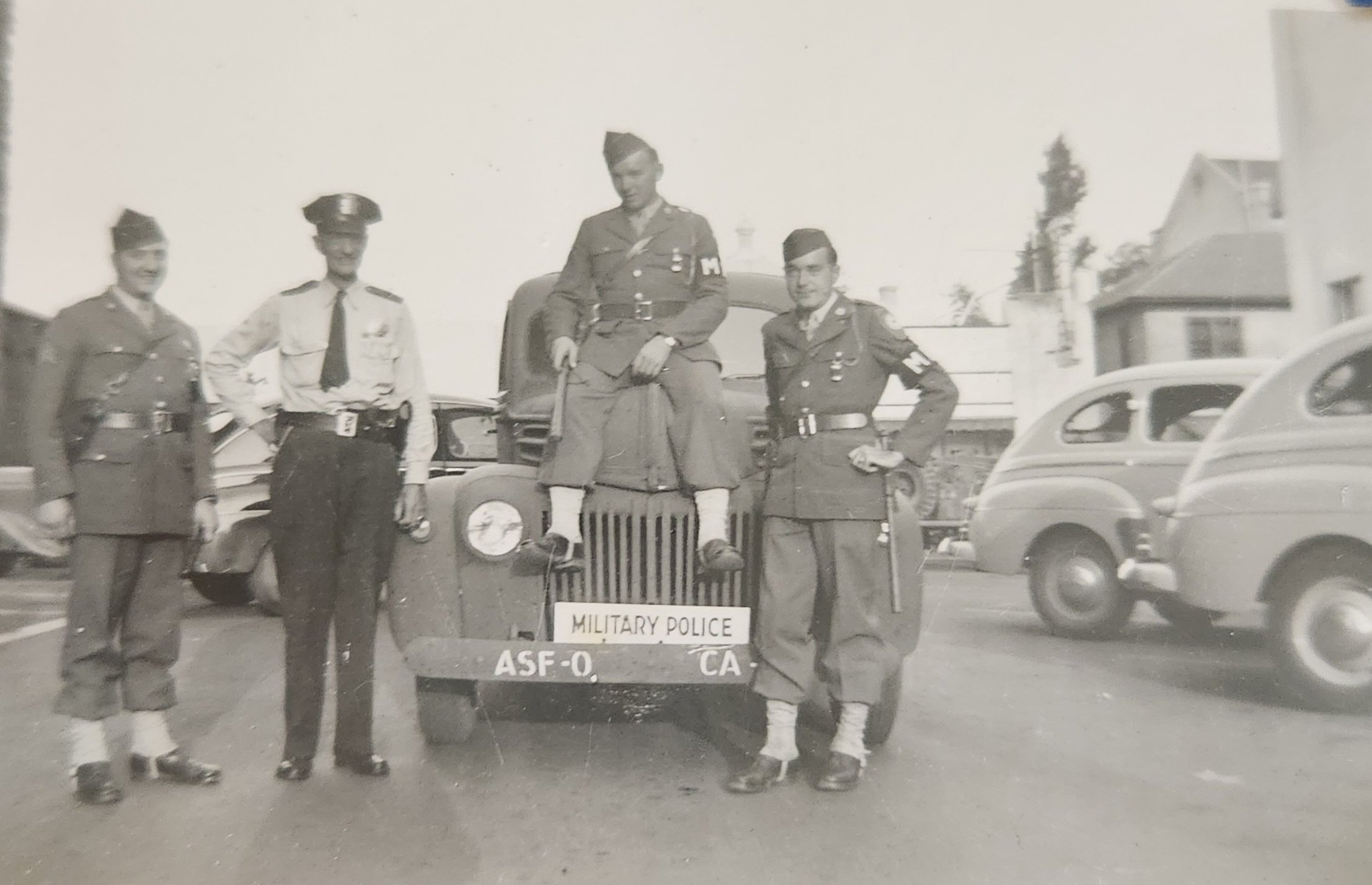 Historical black-and-white photo showing four uniformed military police officers, one sitting on a barrier labeled 'Military Police' and three standing beside him, in an outdoor setting with cars and buildings in the background.